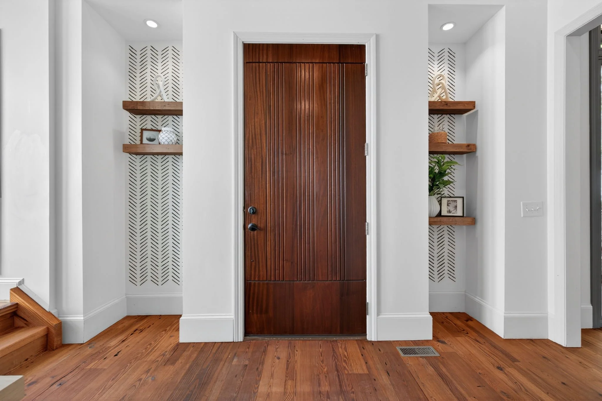 Entryway with a closed wooden door, two floating wooden shelves with decorative items, white walls with black geometric patterns, wooden flooring, and a floor vent.