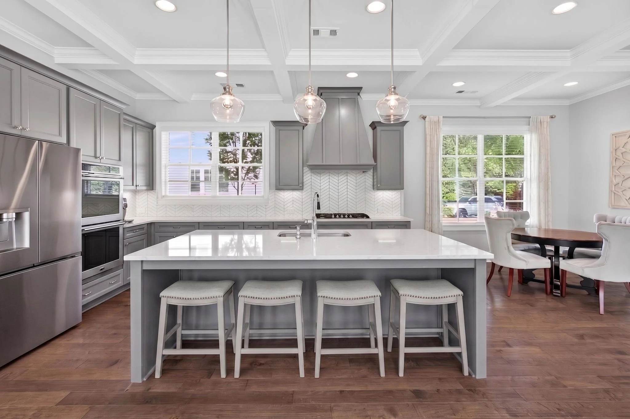 Modern kitchen with gray cabinets, stainless steel appliances, white countertop island, three glass pendant lights, windows with white curtains, wooden dining table with white chairs, hardwood flooring, and a view outside.