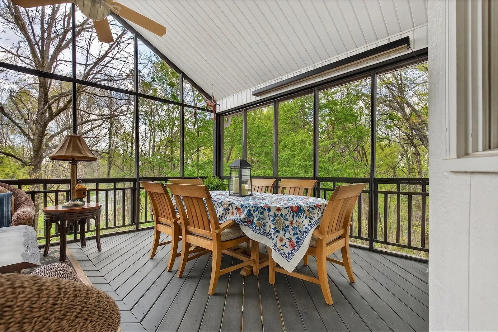  screened porch with wooden dining table, six chairs, decorative tablecloth, lantern, and greenery outside the screens.