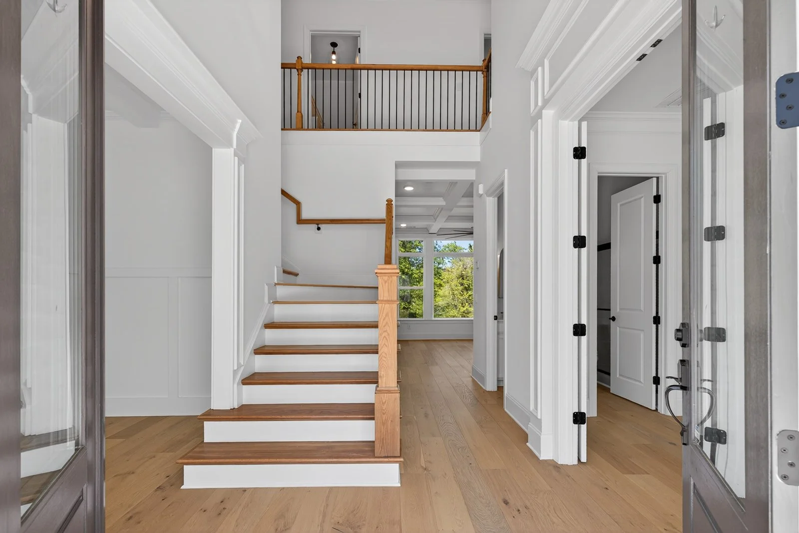 Interior view of a modern house entryway showing a staircase with wooden steps and black handrails, an open closet, and white walls with crown molding, with sunlight coming through large windows in the background.