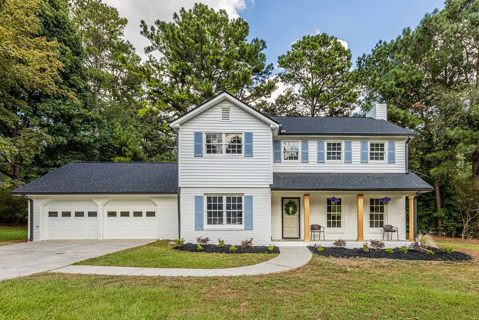 Front view of a two-story white house with blue shutters, a covered porch, a attached garage, and landscaped front yard with walkway, shrubs, and hanging flower baskets, surrounded by trees.