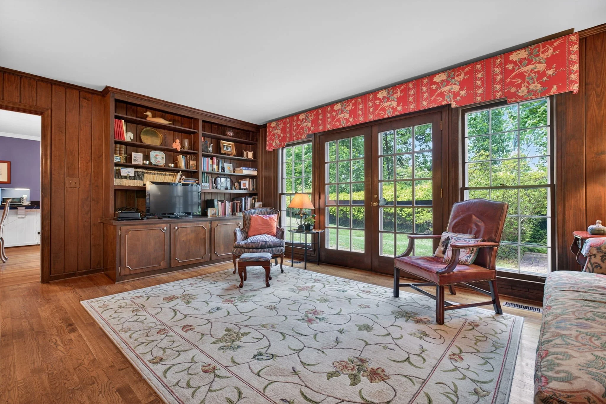 Living room with wooden walls, a large window showing trees outside, a patterned area rug, a leather armchair, and a wooden bookshelf with decorative items and books.