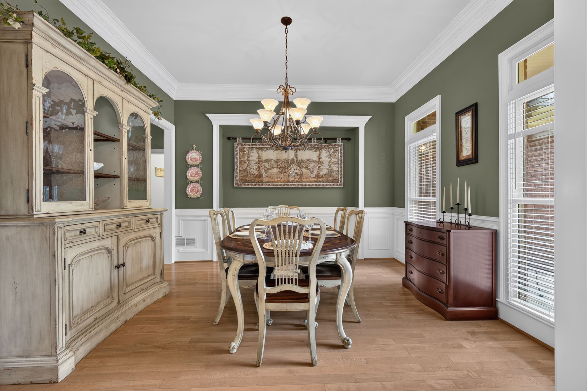 Dining room with green walls, white trim, a chandelier, large windows with blinds, a wooden table with chairs, a distressed cream-colored cabinet, and a dark wood dresser with candleholders.