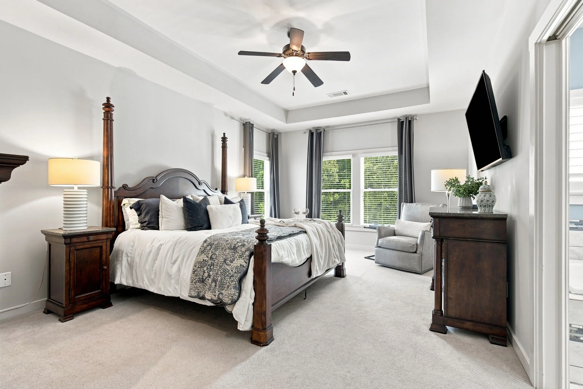 Bedroom with large bed, wooden headboard, bedside lamps, gray curtains, flat-screen TV on wall, white armchair, and decorated dresser near window.