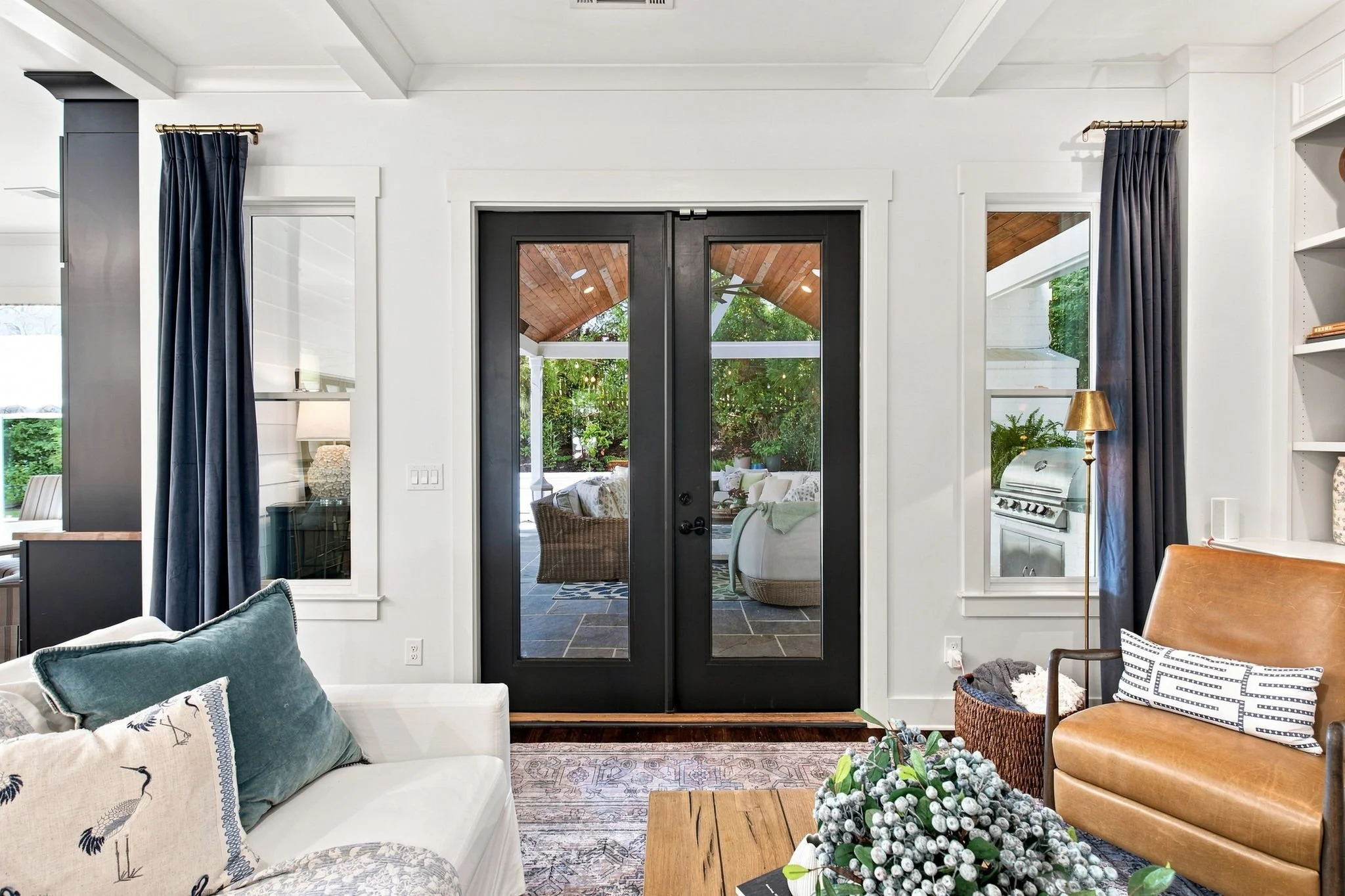 Living room with white walls, dark wood framed glass door leading outside, blue curtains, cream sofa with decorative pillows, wooden coffee table with flower arrangement, tan leather armchair, adjacent kitchen with a silver grill outside, cozy outdoo