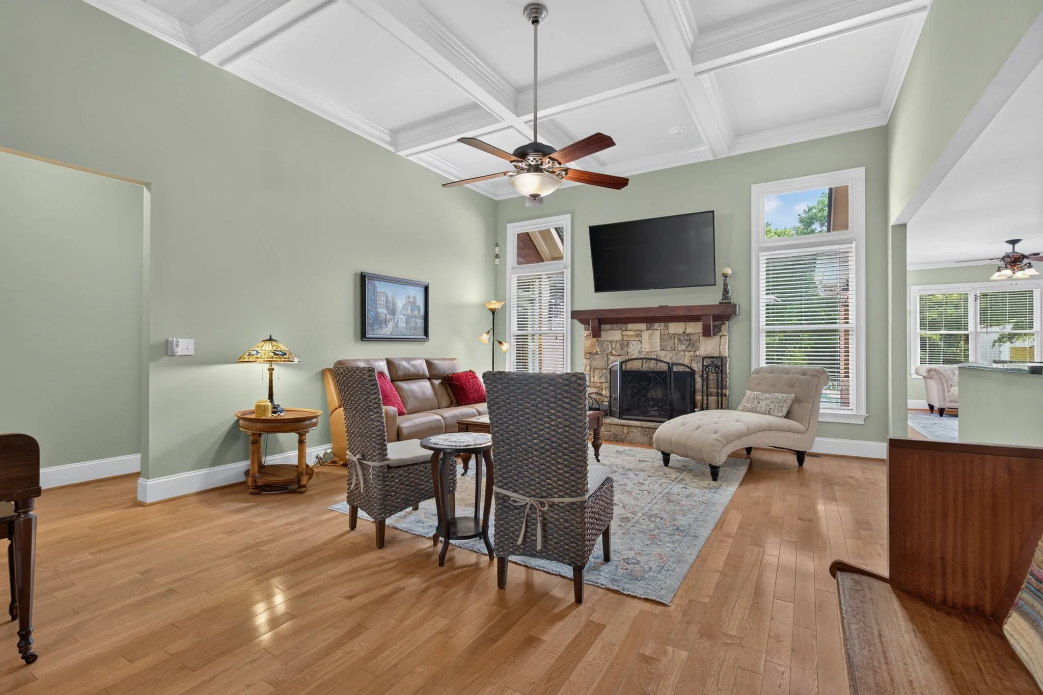 A living room with green walls, large windows with shades, a stone fireplace with a wooden mantel, sofas, a coffee table, a TV mounted above the fireplace, and a ceiling fan.