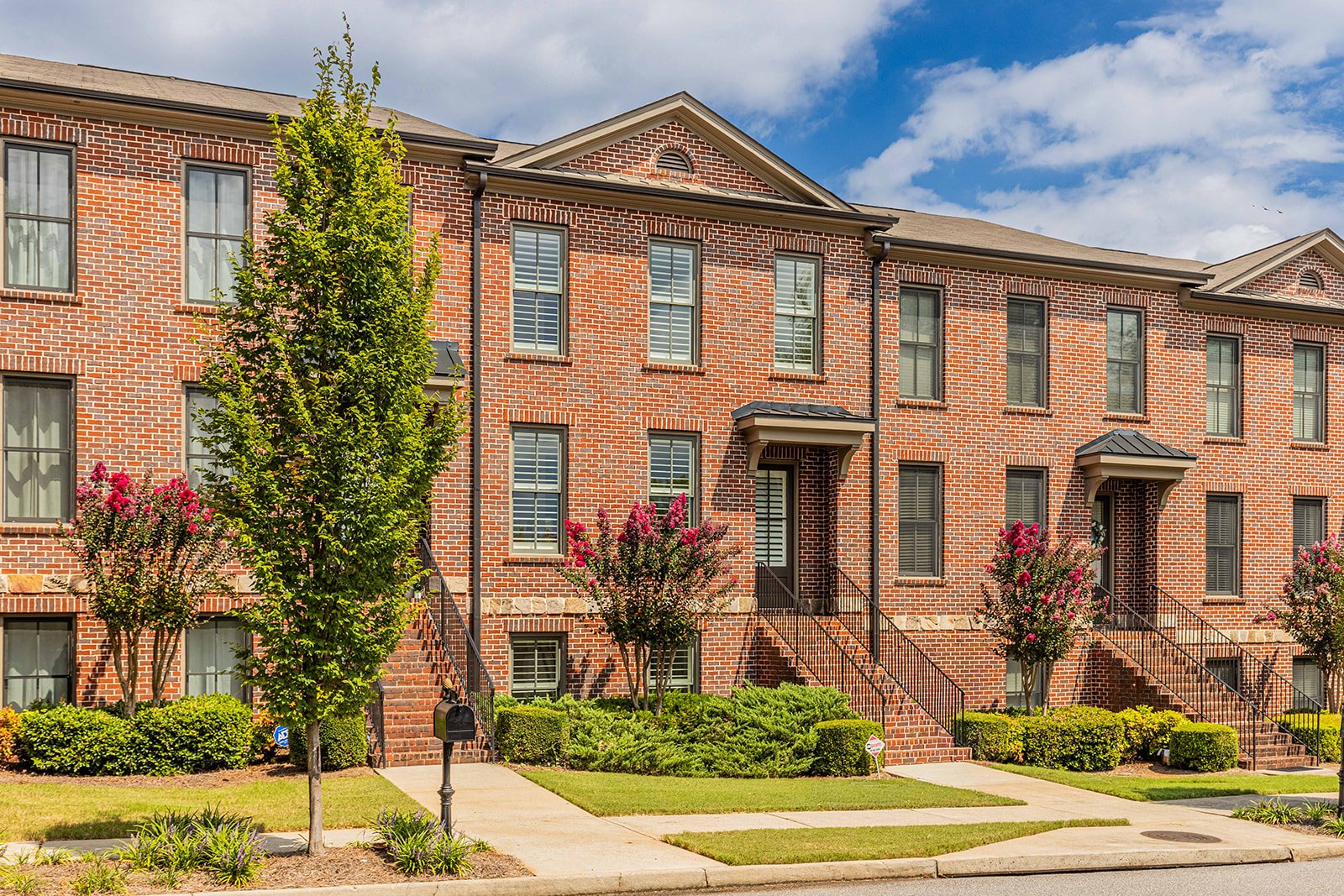 A row of brick townhouses with front steps, green lawns, flowering trees, and a sidewalk under a partly cloudy sky.