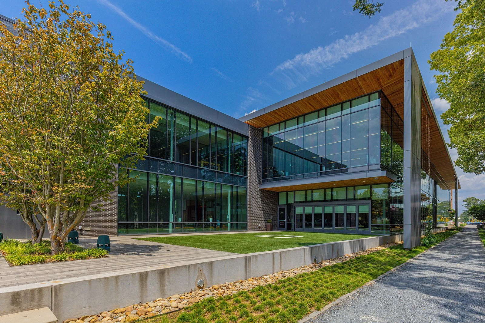Modern office building with large glass windows, wooden accents, surrounded by trees, with a concrete pathway and neatly maintained grass.