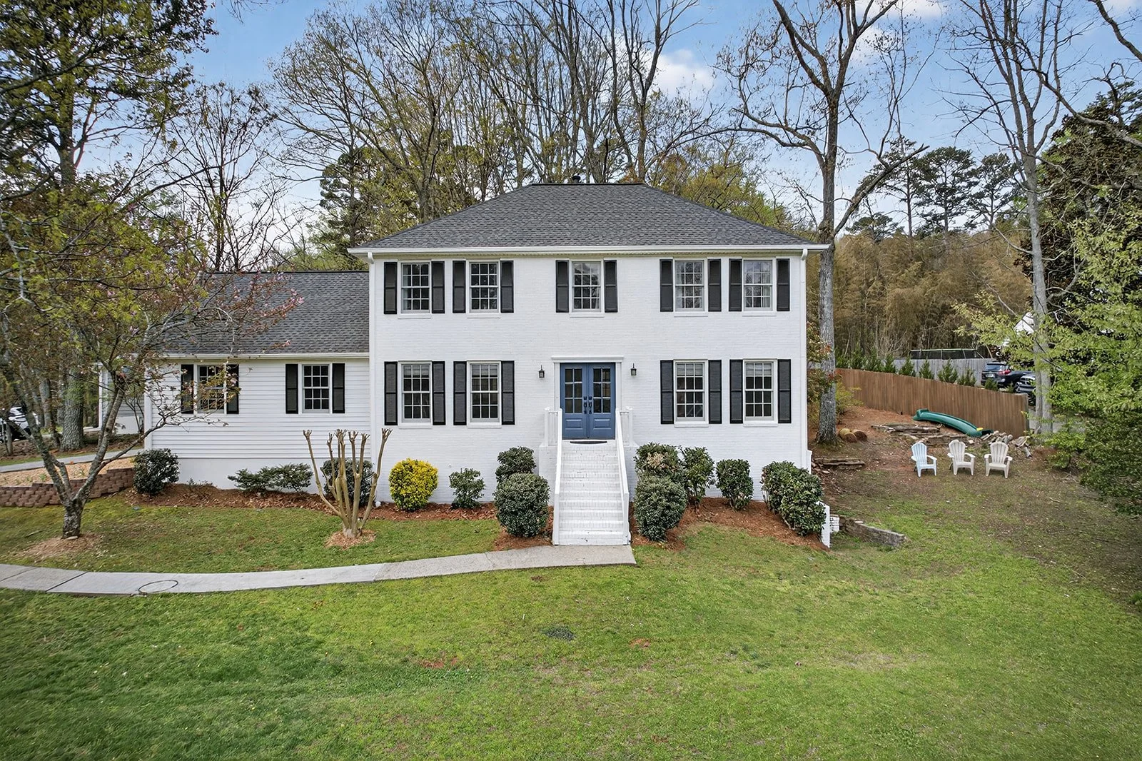 Front view of a two-story white house with black shutters, blue front door, and front stairs, surrounded by green lawn and trees, with outdoor chairs and a slide visible on the backyard hillside.