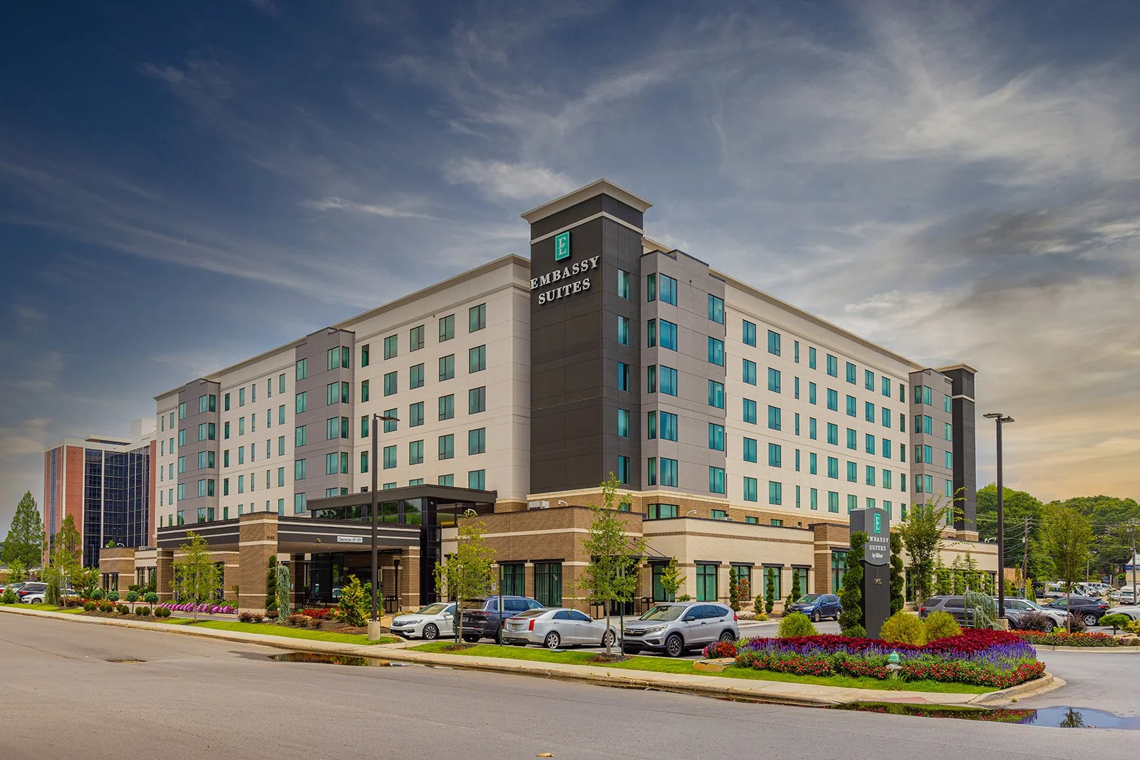 A modern multi-story hotel building labeled 'Embassy Suites' with a parking lot and landscaped area featuring flowers and small trees in front.
