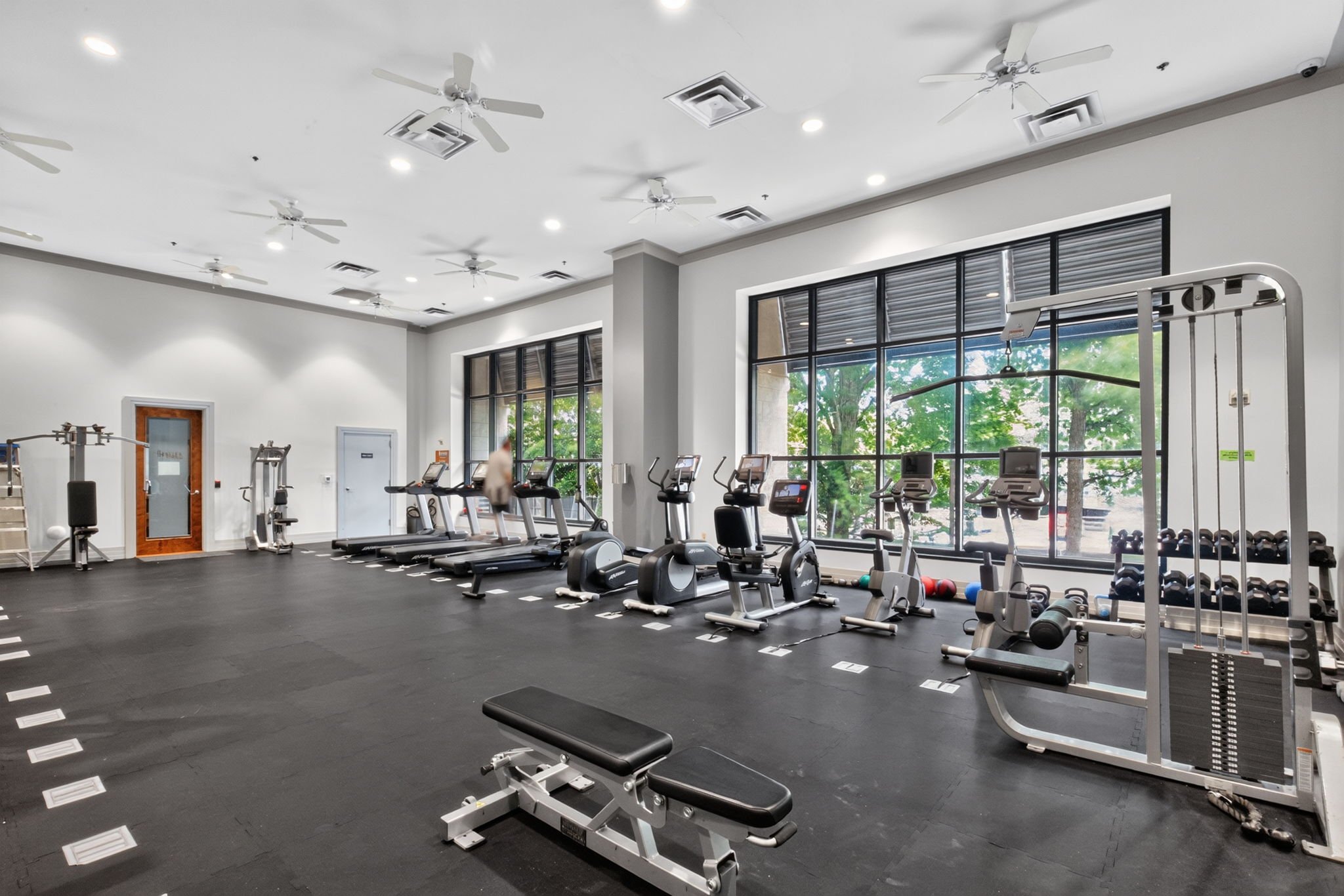 Interior of a modern gym with cardio equipment, weight racks, large windows, ceiling fans, and ceiling vents.