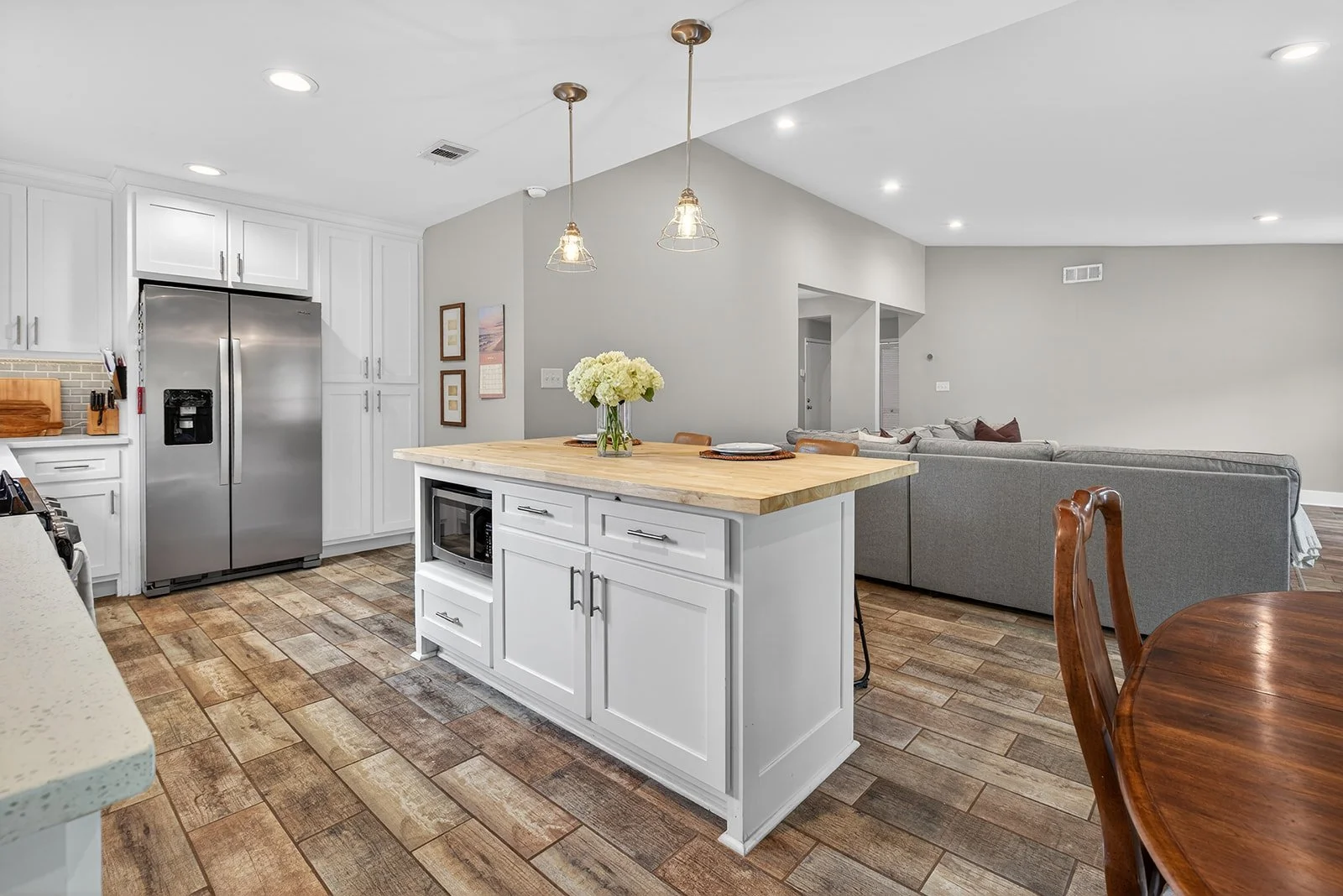 Open-plan kitchen and living room with white cabinetry, stainless steel refrigerator, wooden kitchen island with a vase of flowers, gray sofa, wood dining table, and wood plank flooring.
