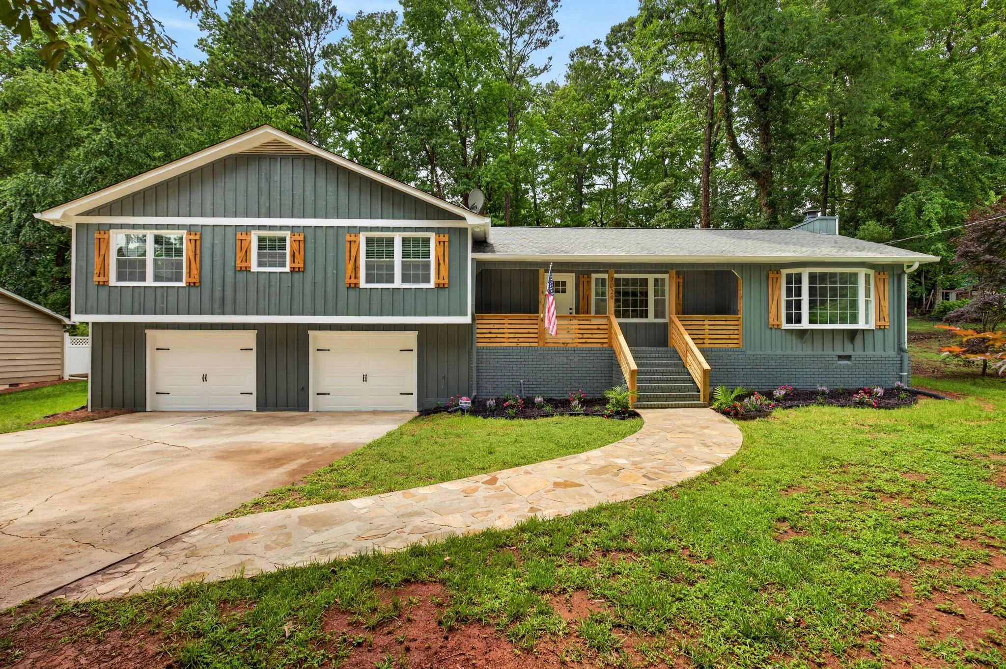 A two-story house painted gray with white trim and wooden shutters, featuring a front porch with wooden stairs, a flag, and surrounded by a green lawn and trees.