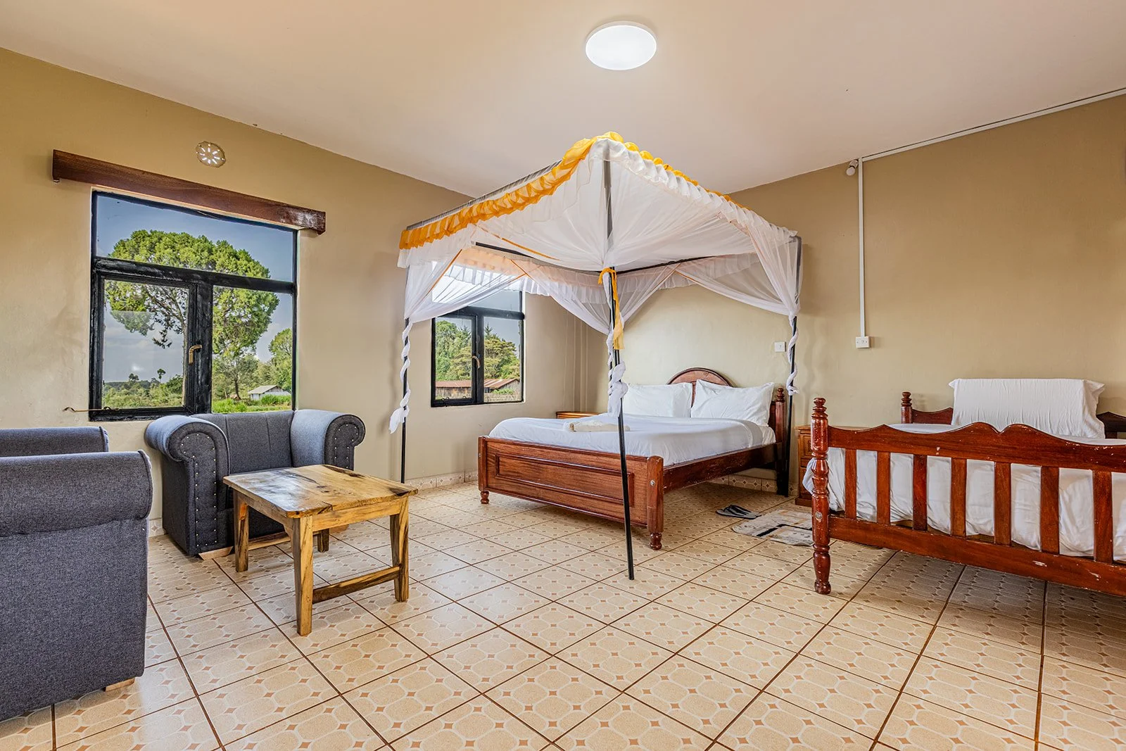A room with two beds, one with a canopy, a small table, and two armchairs near a window with a view of trees outside.