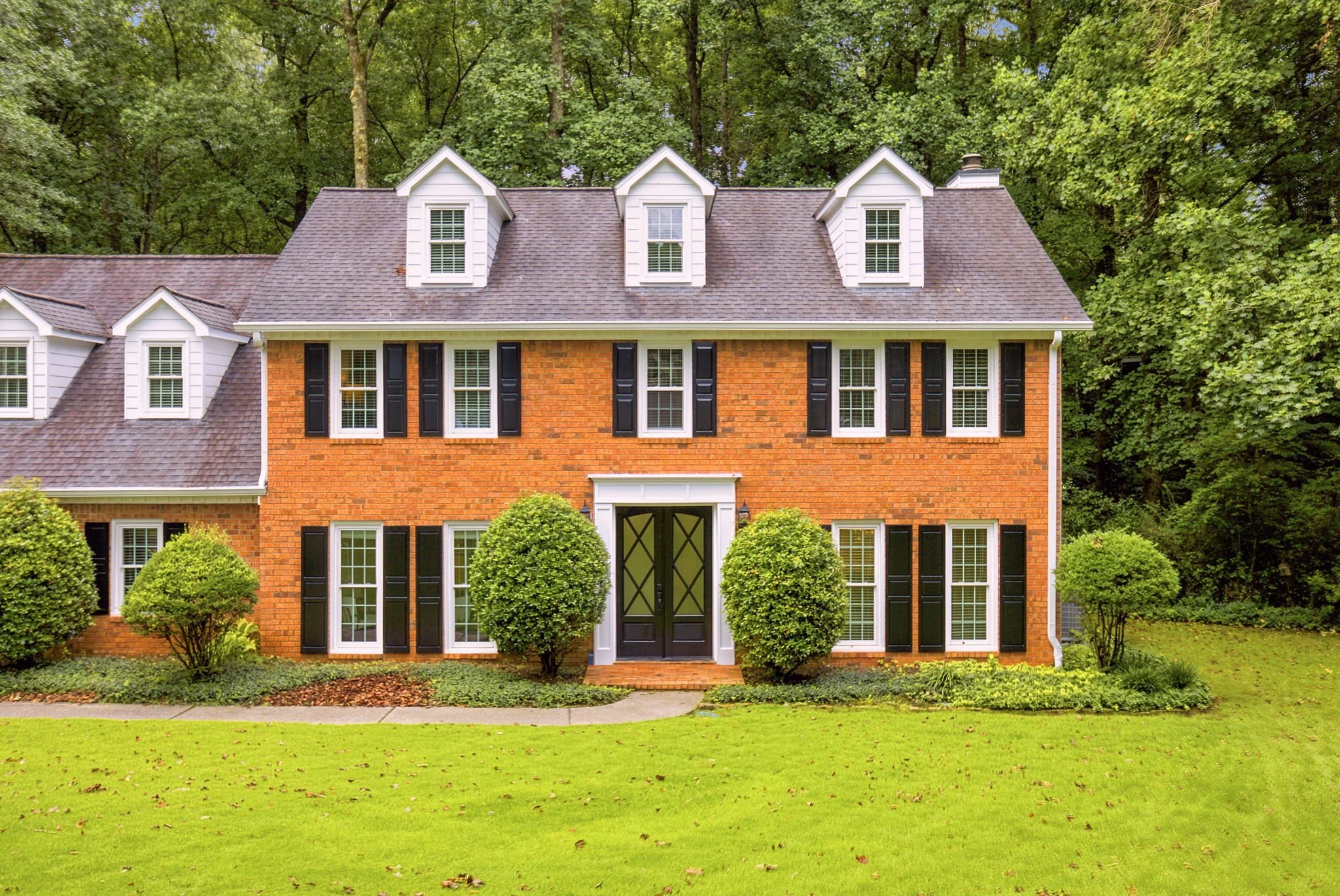 A brick house with black shutters, multiple white-framed windows, a black front door, and a landscaped front yard with green bushes, surrounded by tall trees.