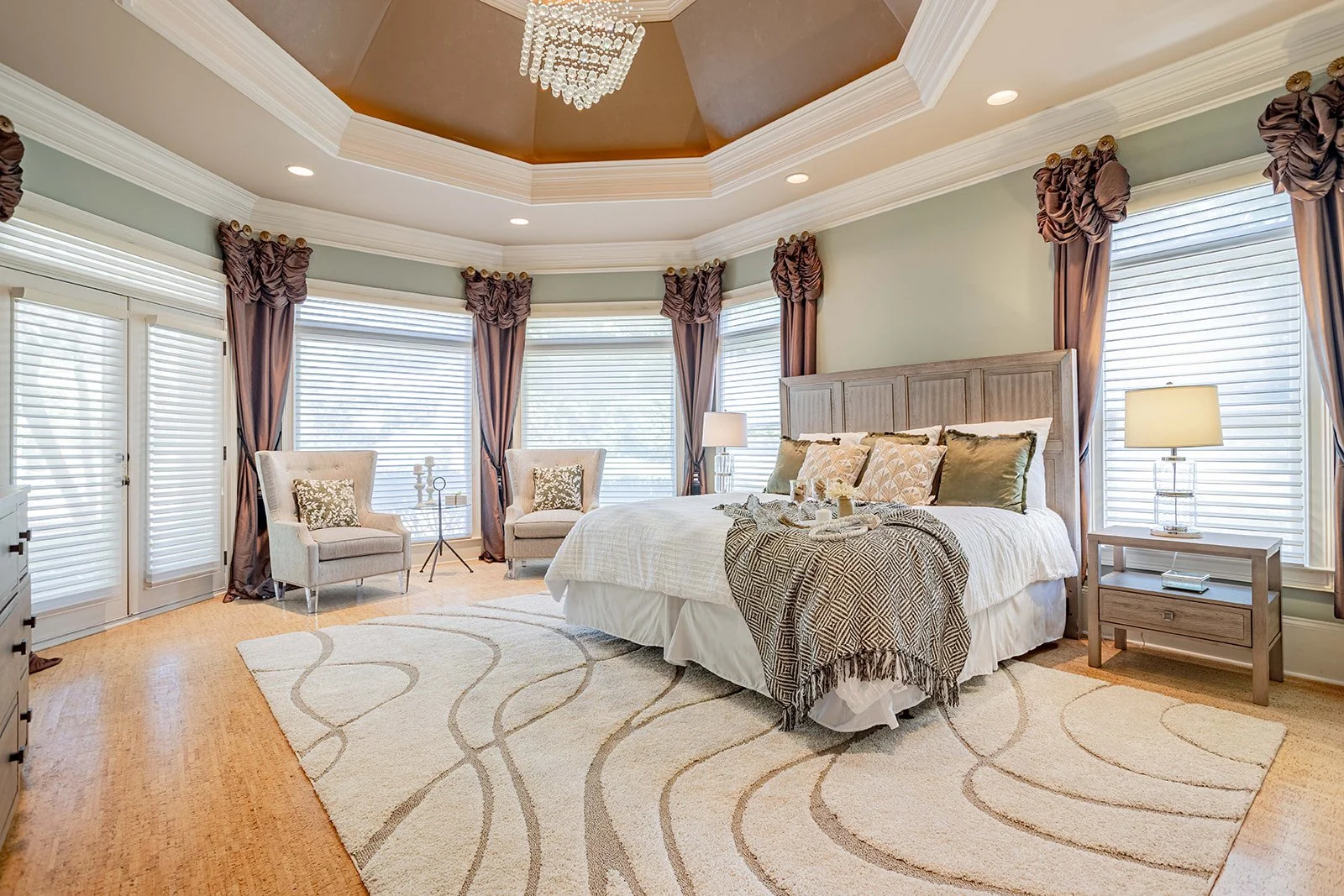 A bedroom with a large bed, window seating with two armchairs, and a window with blinds and curtains, decorated in soft neutral tones.