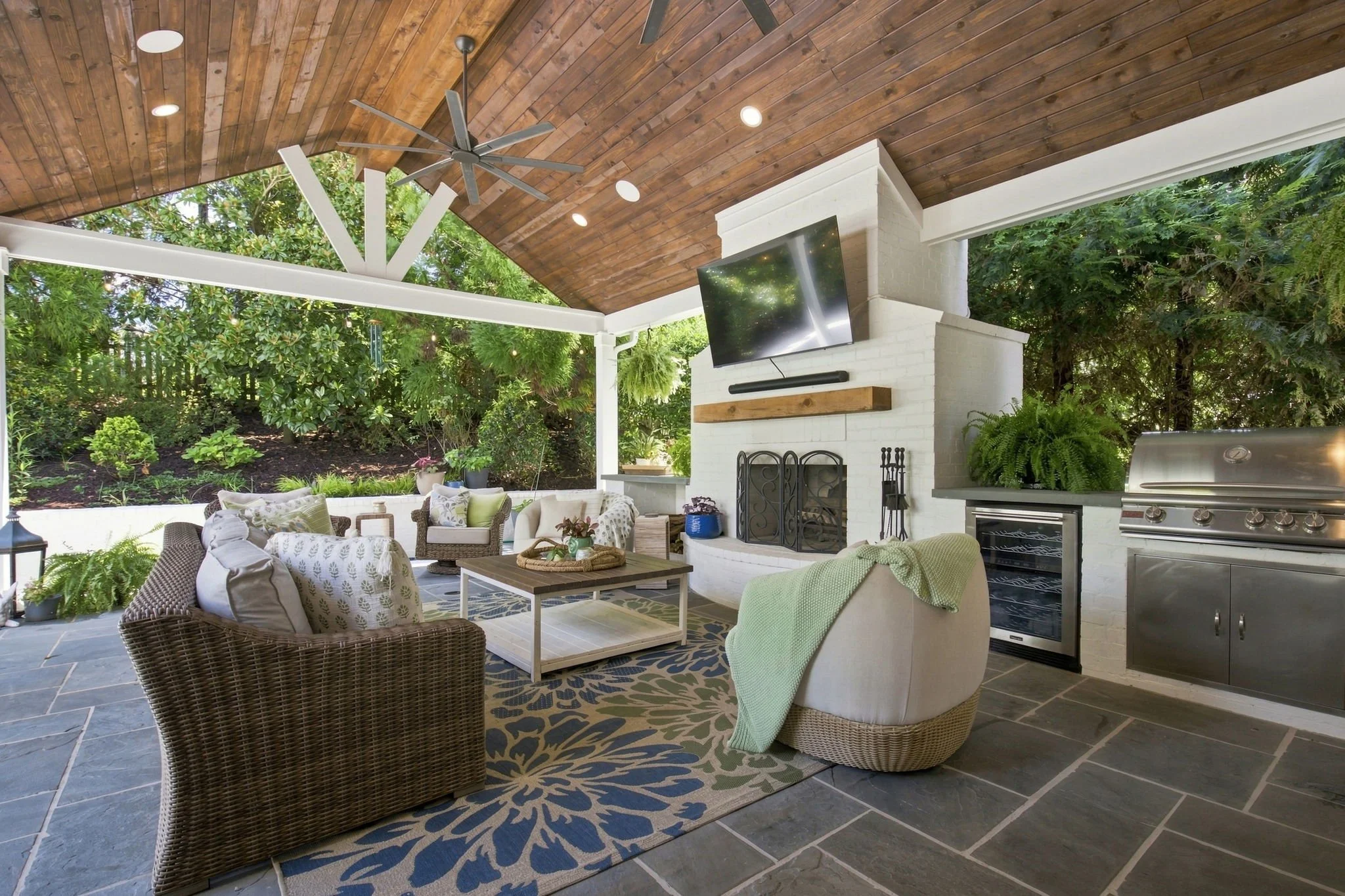 A covered outdoor patio with woven wicker furniture, a rug, a fireplace, a TV, and a ceiling fan, surrounded by lush greenery.