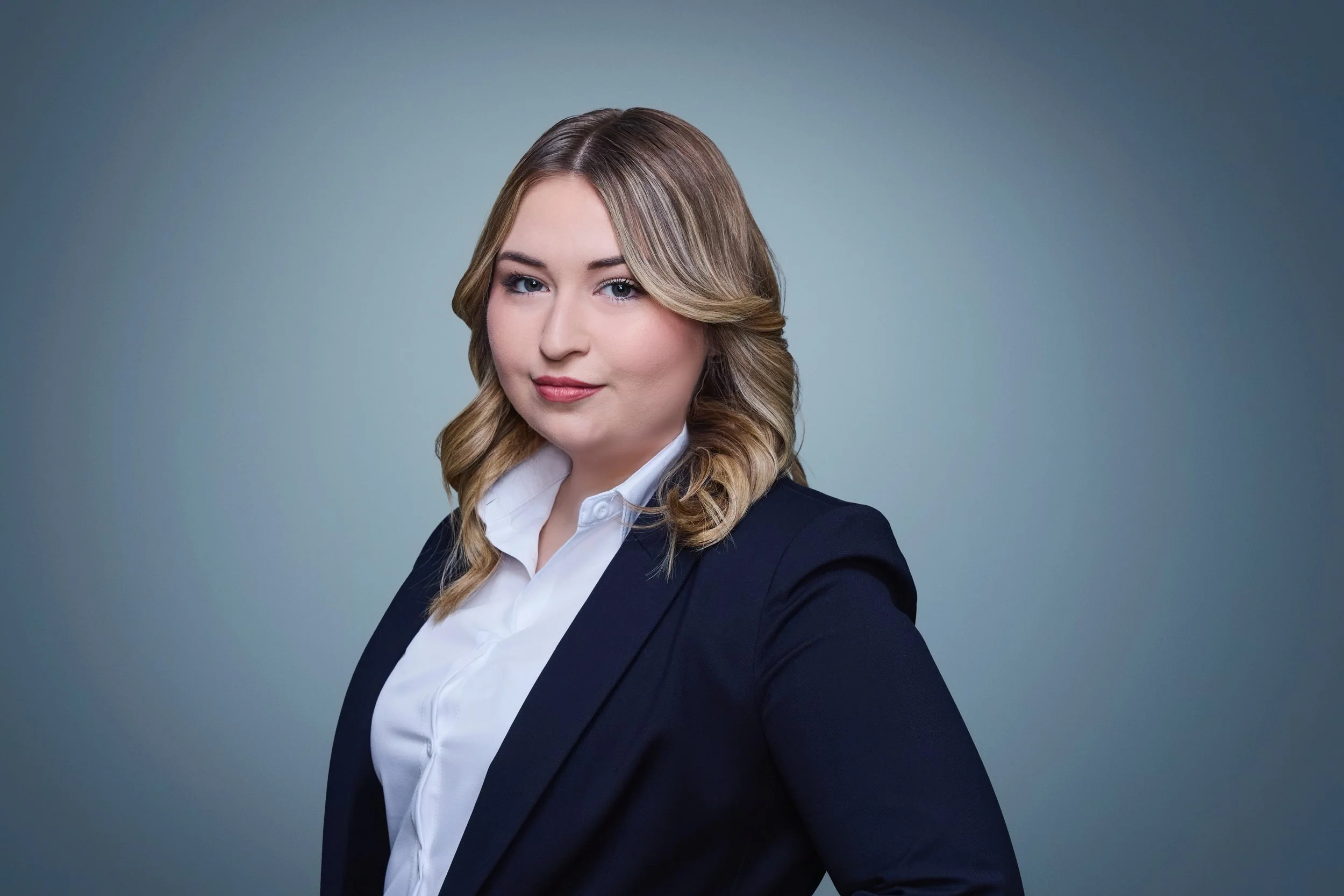 A young woman with blonde hair styled in loose curls, wearing a dark blazer and white shirt, posing against a plain blue-gray background.
