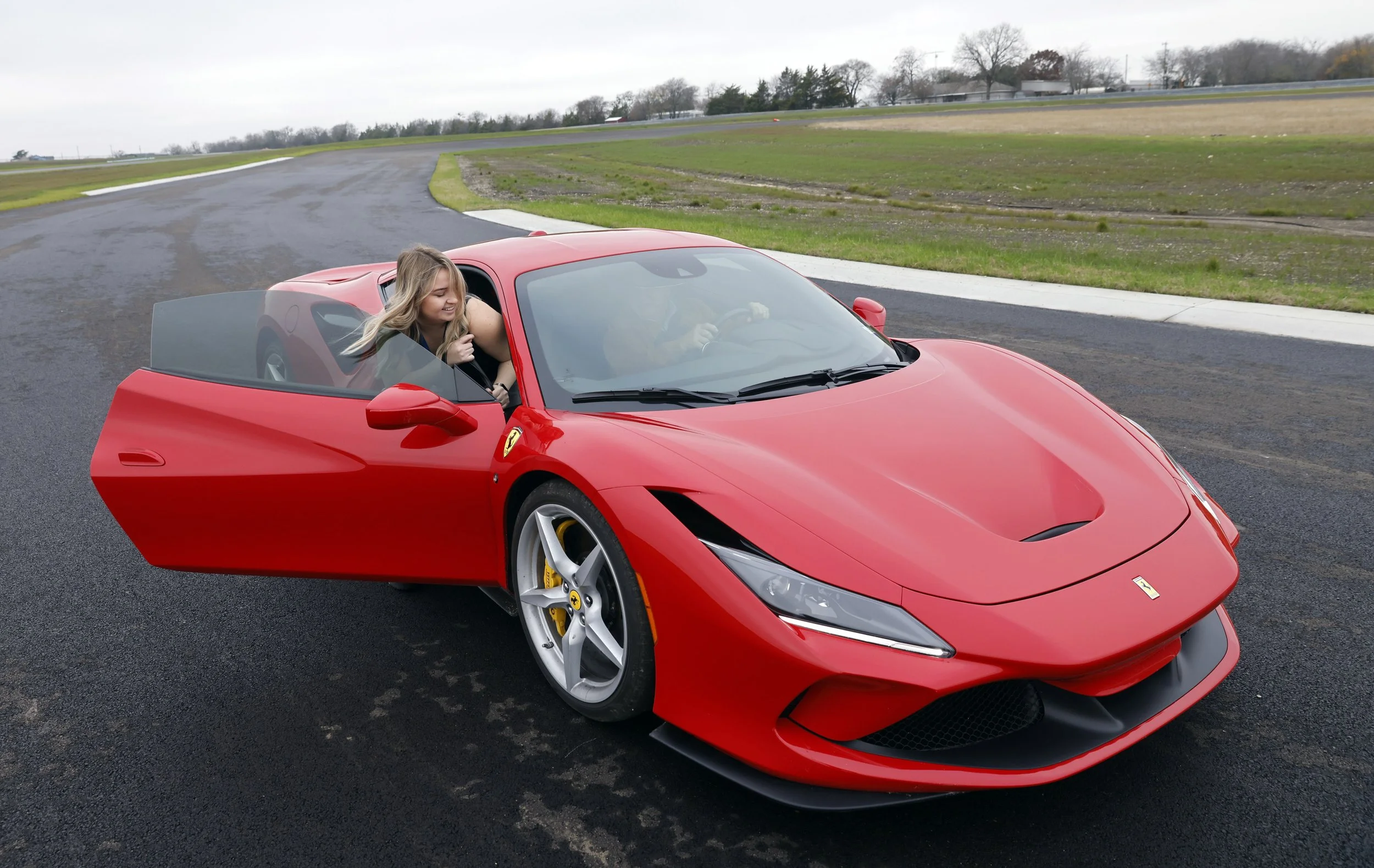 Alexandra Skores leaning out of a red sports car, smiling, on a race track with a curved asphalt track and grassy areas in the background.