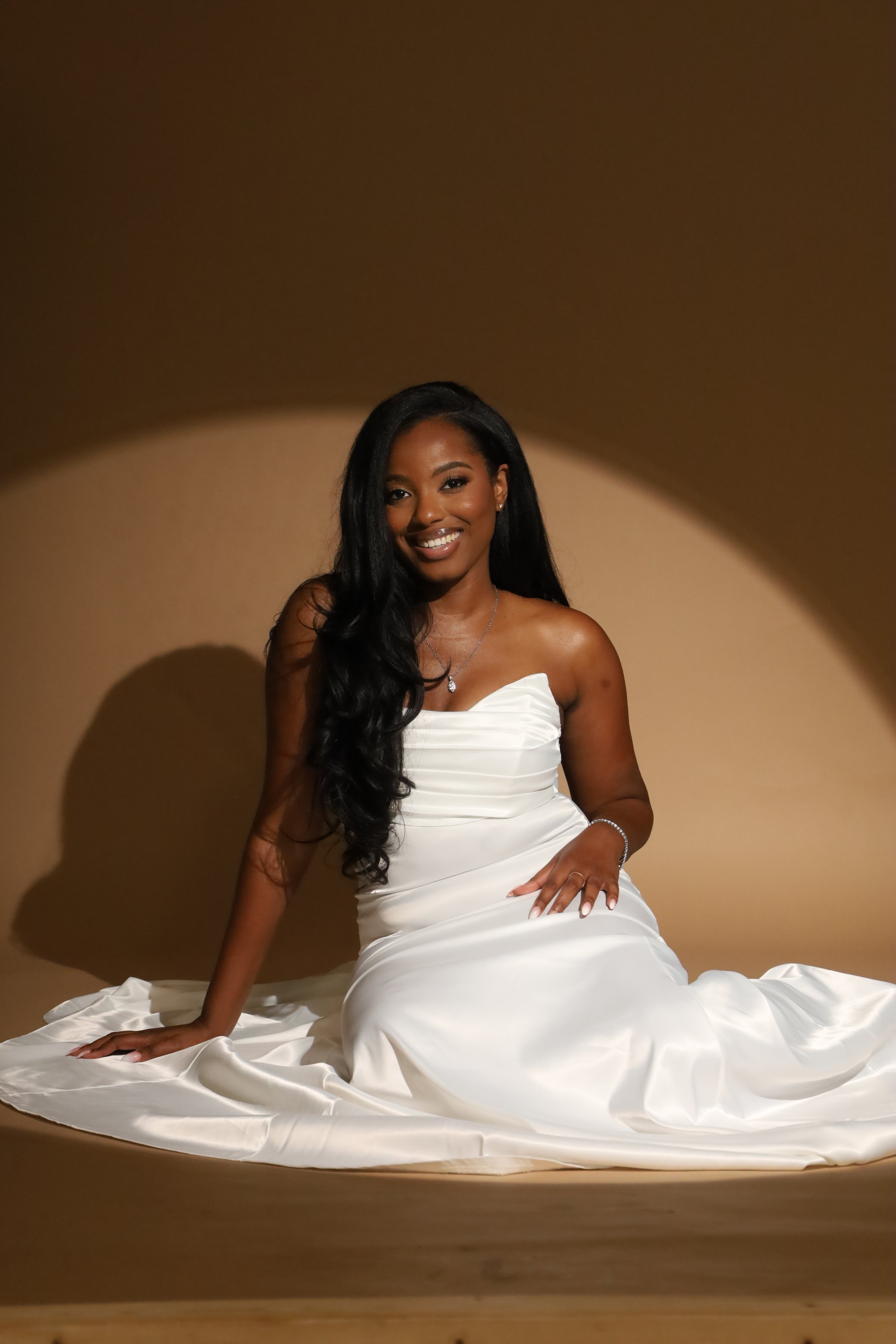A woman in a white satin dress sitting on the floor with one hand on the ground and the other resting on her knee, smiling against a beige background with an oval-shaped shadow.