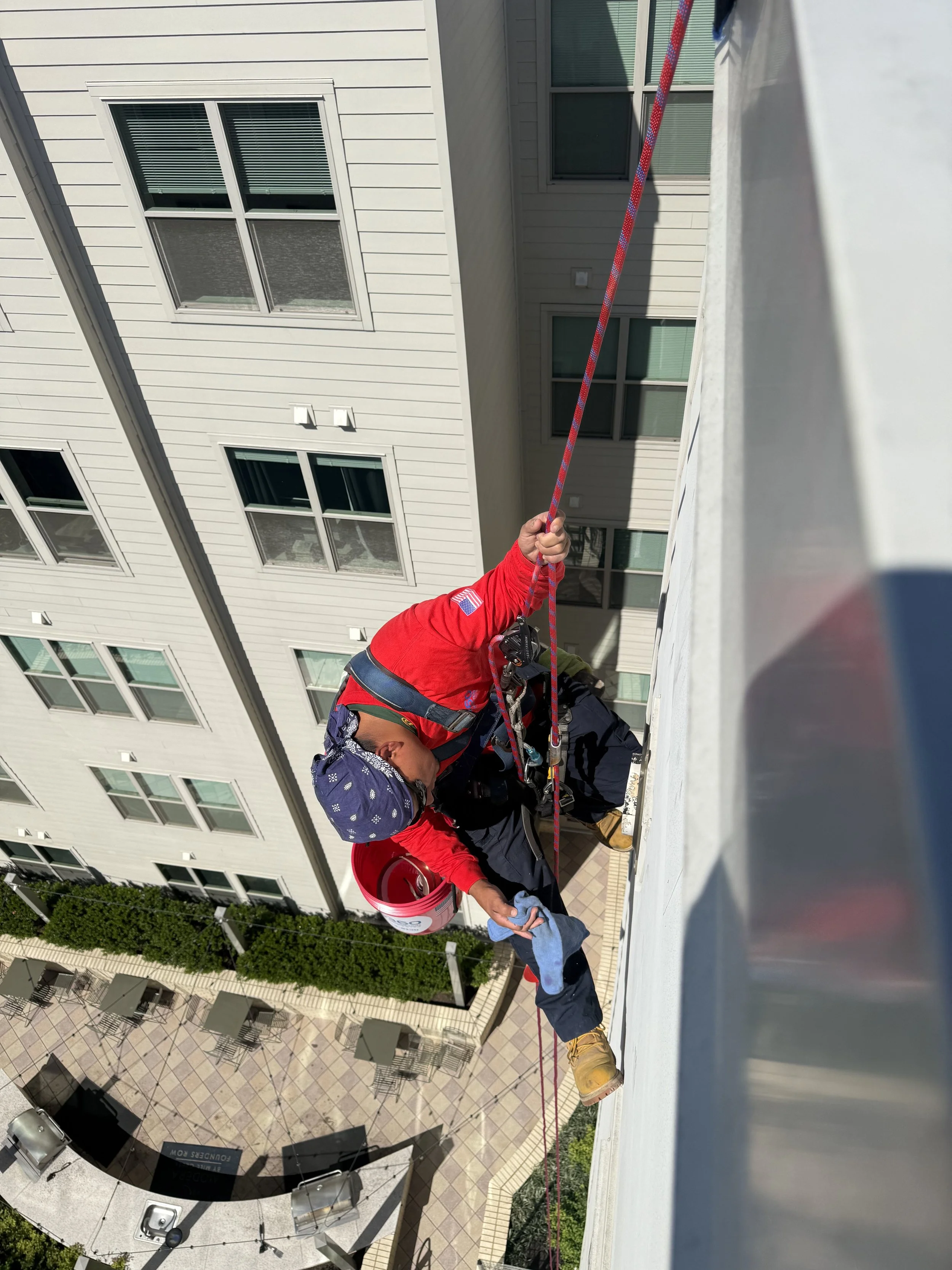 Worker in red jacket and helmet cleaning the exterior of a tall building using a harness and safety rope, with a view of other buildings and a patio below.