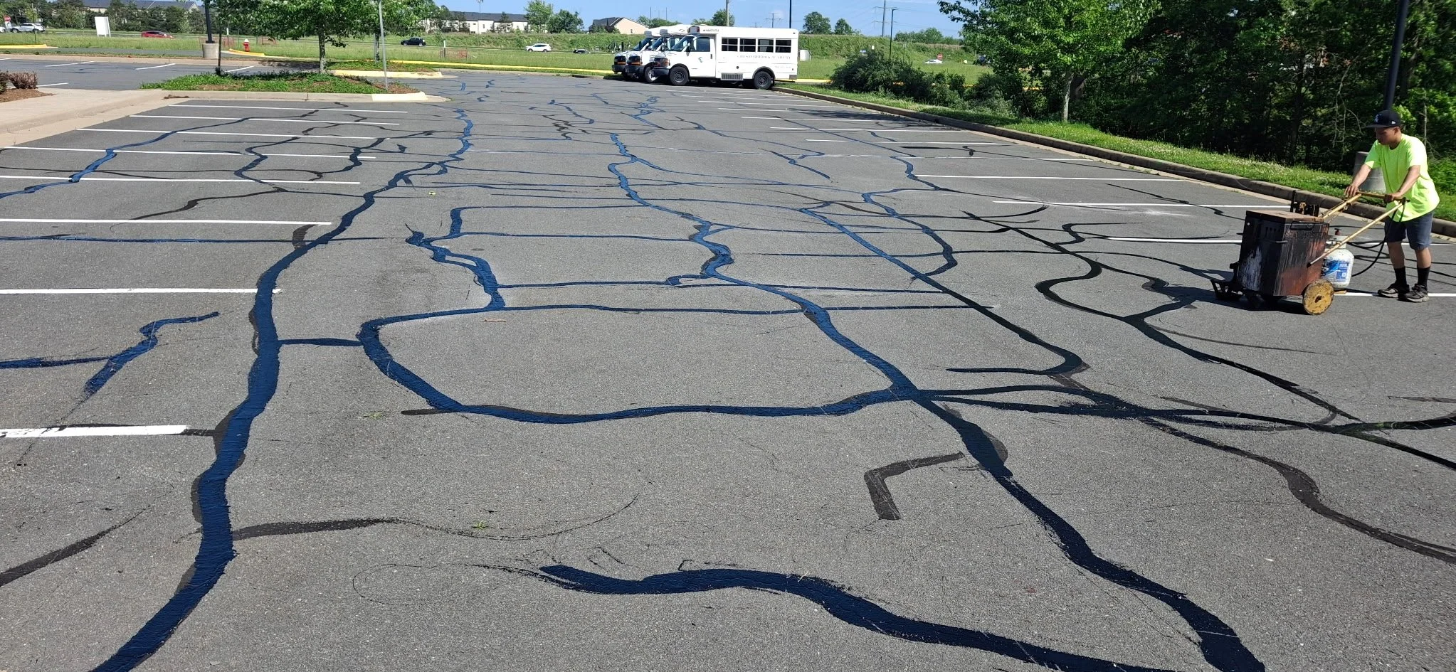 A person in a neon green shirt and black shorts is sealing cracks in an empty parking lot with a hot rubberized crack sealant machine. There are multiple black and blue cracks repaired on the asphalt surface, with some white parking lines visible. In the background, there is a white bus, some trees, a grassy area, and a few buildings under a clear blue sky.