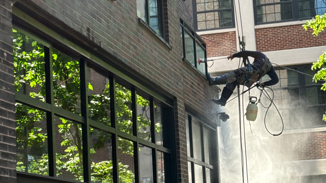 Worker wearing safety gear using a harness and rope to clean or repair the exterior window of a multi-story brick building, with dust or steam visible in the air.