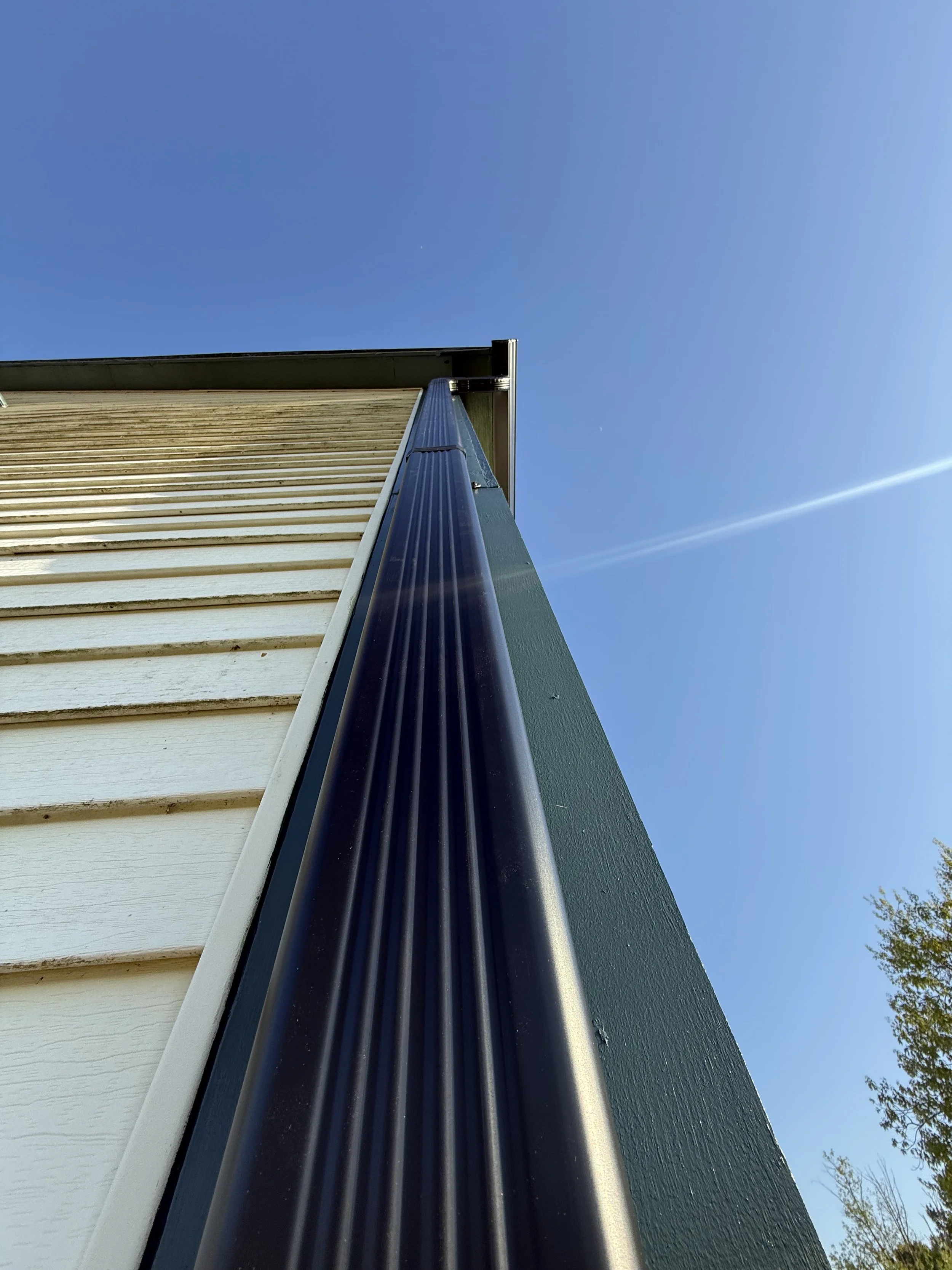 A close-up, upward view of a house's gutter and siding against a clear blue sky with a faint contrail.