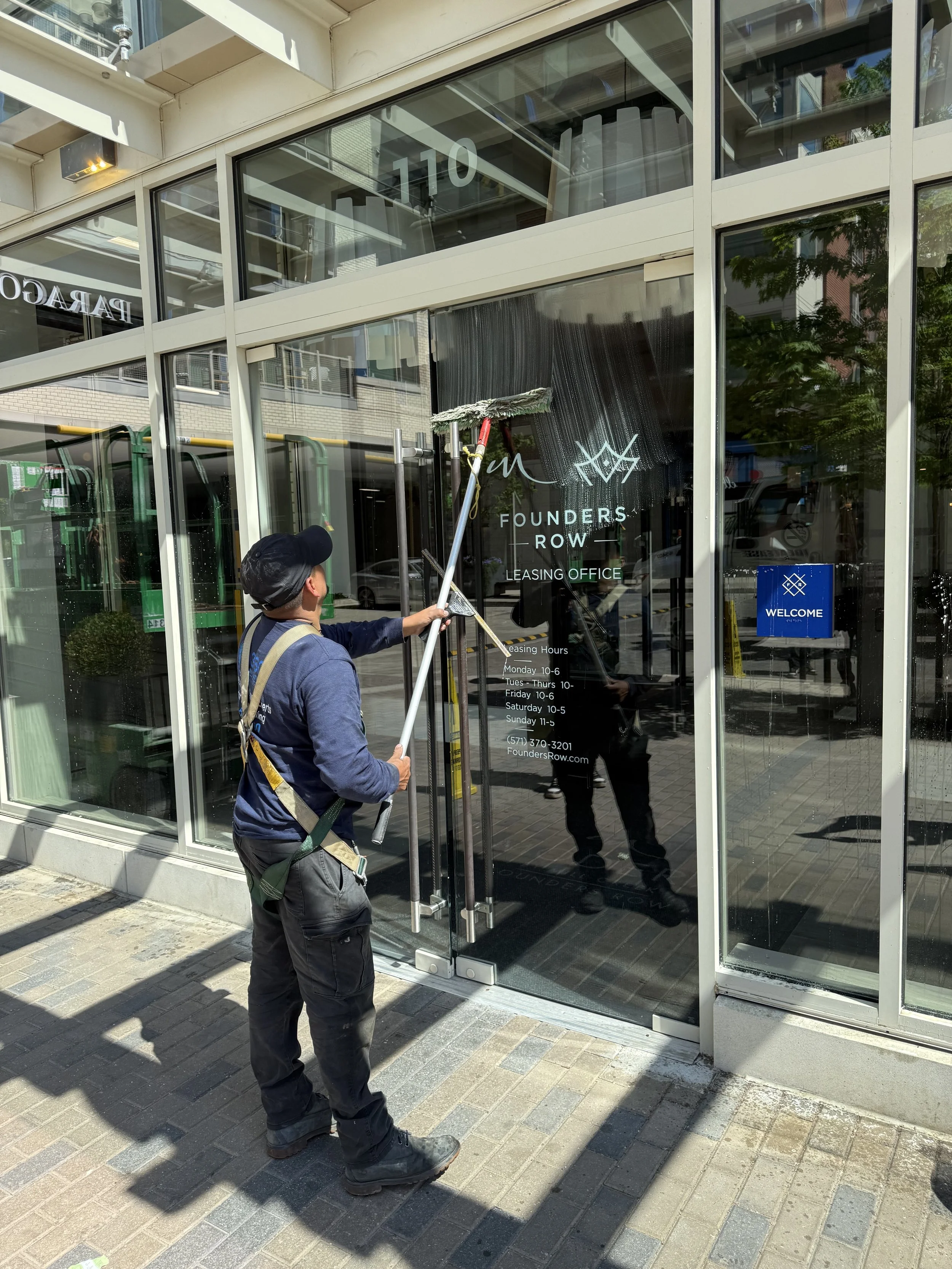 A man cleaning the glass exterior of a commercial building with a squeegee. The building has large glass windows and a door with signage for Founders Row leasing office. The street and nearby buildings are reflected in the glass.