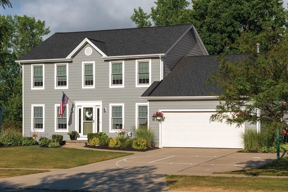 A two-story gray house with white trim, a black roof, and multiple windows. The front door has a wreath, and there is an American flag next to it. The house has a two-car garage, and the driveway has a basketball court marking. The front yard is land