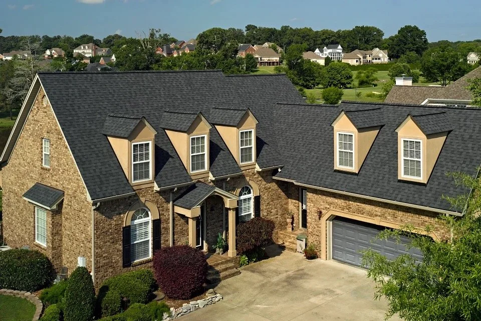 A large brick house with a dark shingle roof and multiple dormer windows, surrounded by well-maintained bushes and trees, with a concrete driveway leading to a two-car garage.