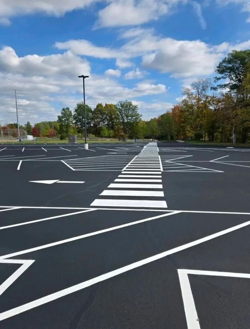Empty parking lot with striped crosswalk and painted parking space lines, surrounded by trees with autumn foliage and a partly cloudy sky.