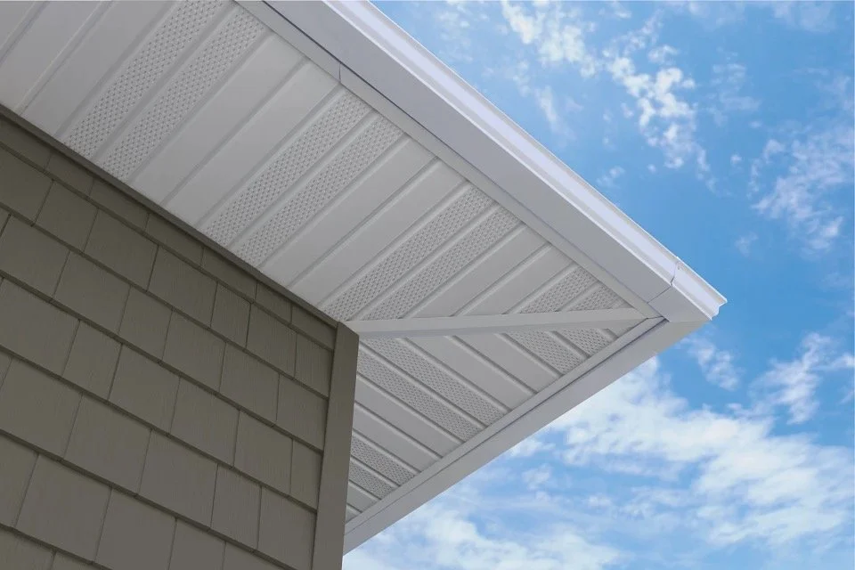Upward view of house eaves showing white vinyl soffit and beige shingle siding against a blue sky with scattered clouds.