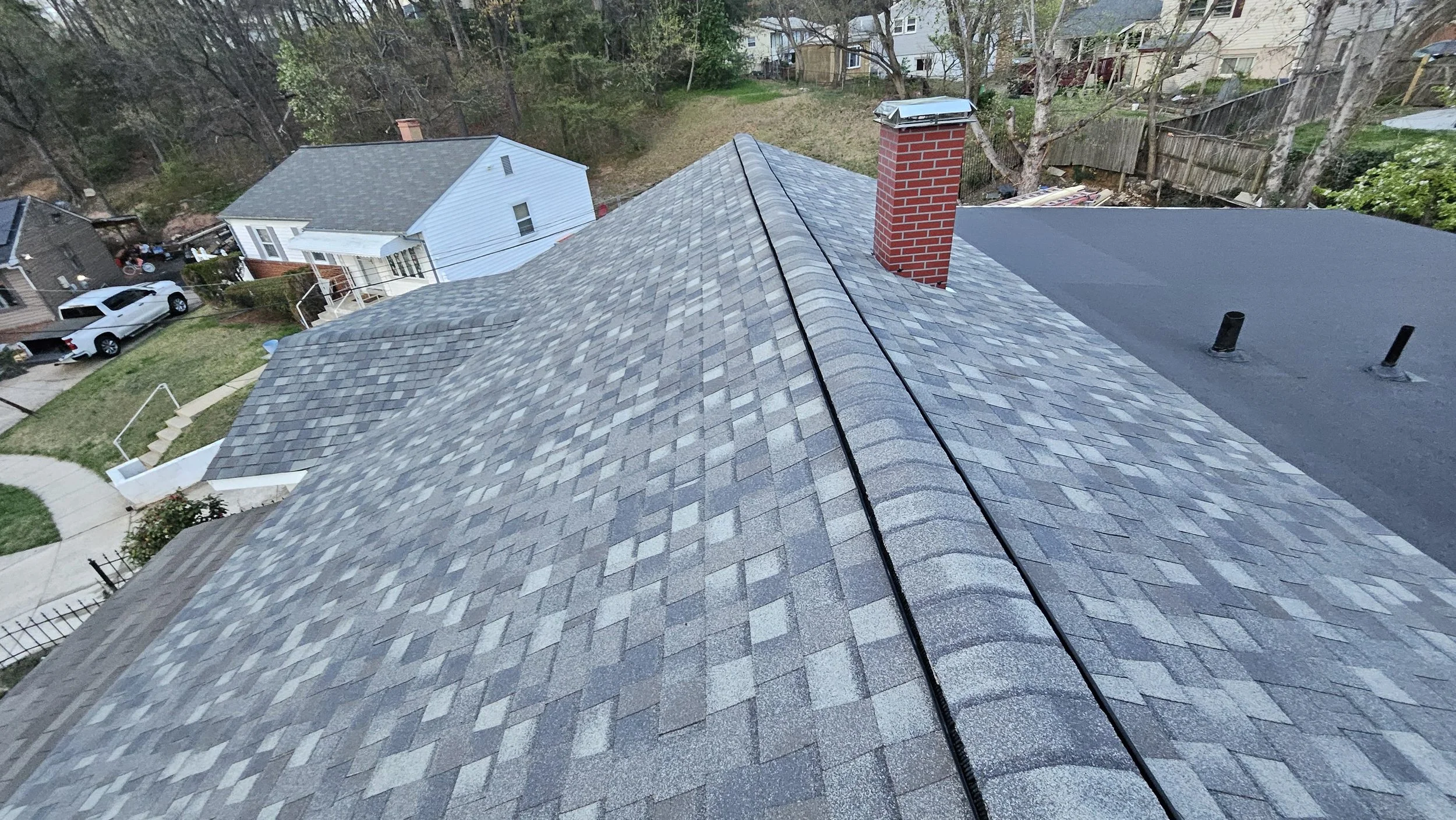 Aerial view of rooftops with gray shingles, brick chimney, and surrounding houses, trees, and yard.