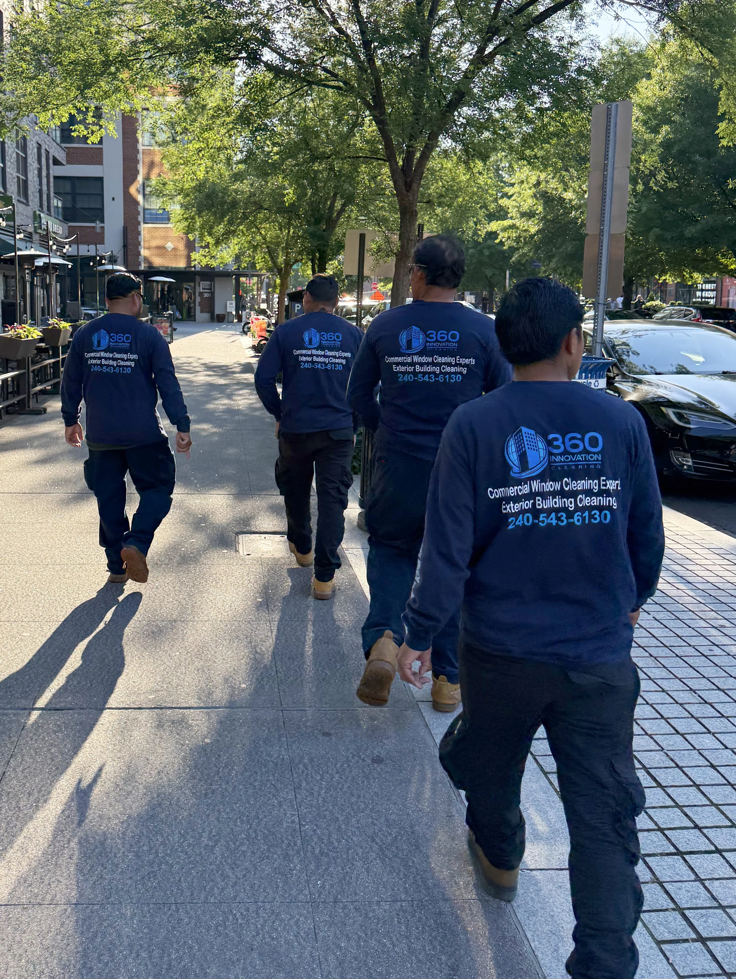 A group of workers walking on a city sidewalk, wearing matching uniforms that say '360 Innovation' and offer exterior building cleaning services, with trees, parked cars, and buildings in the background.