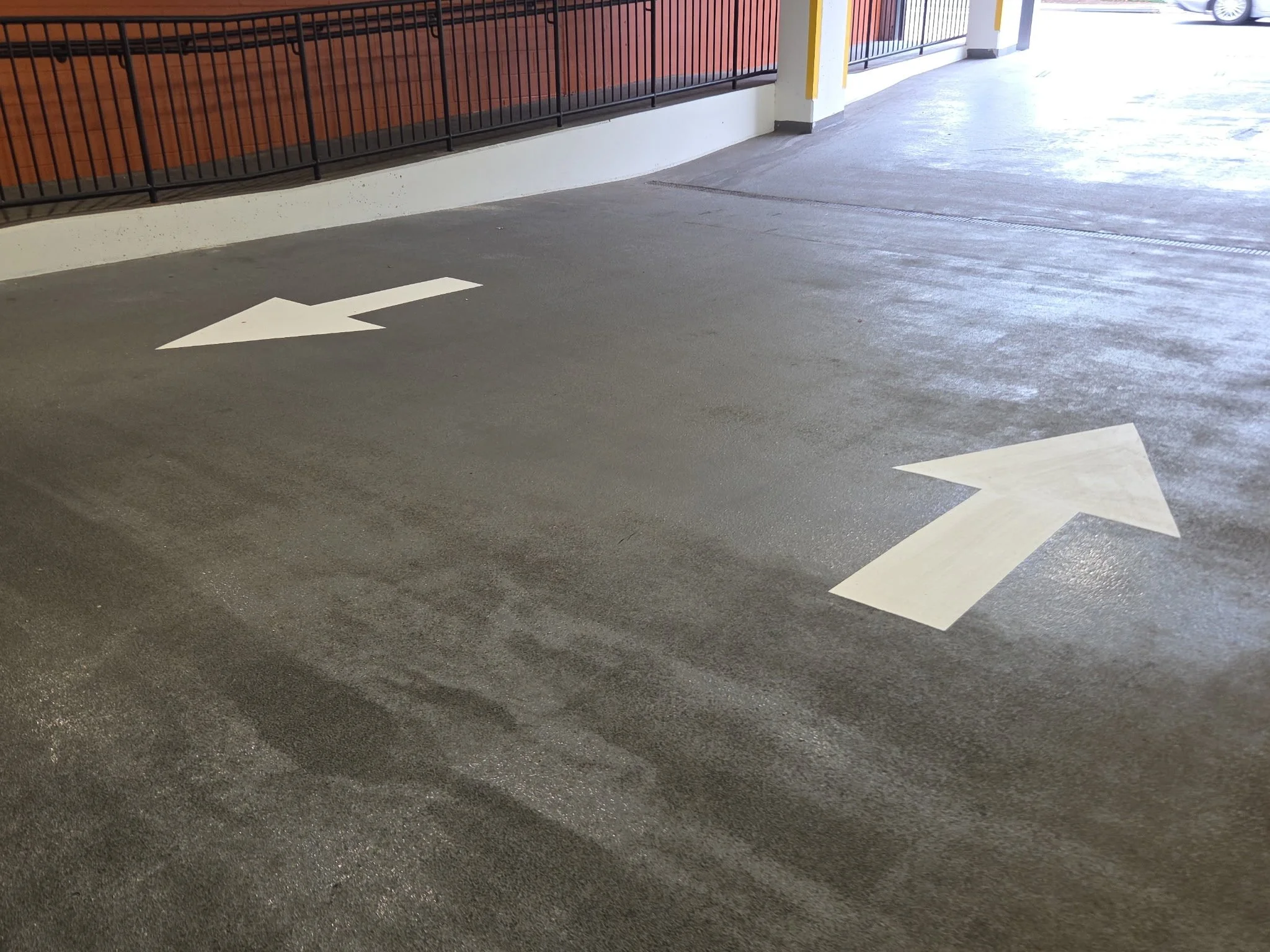 A parking lot with white directional arrows painted on the dark asphalt ground, adjacent to a building wall with a railing.