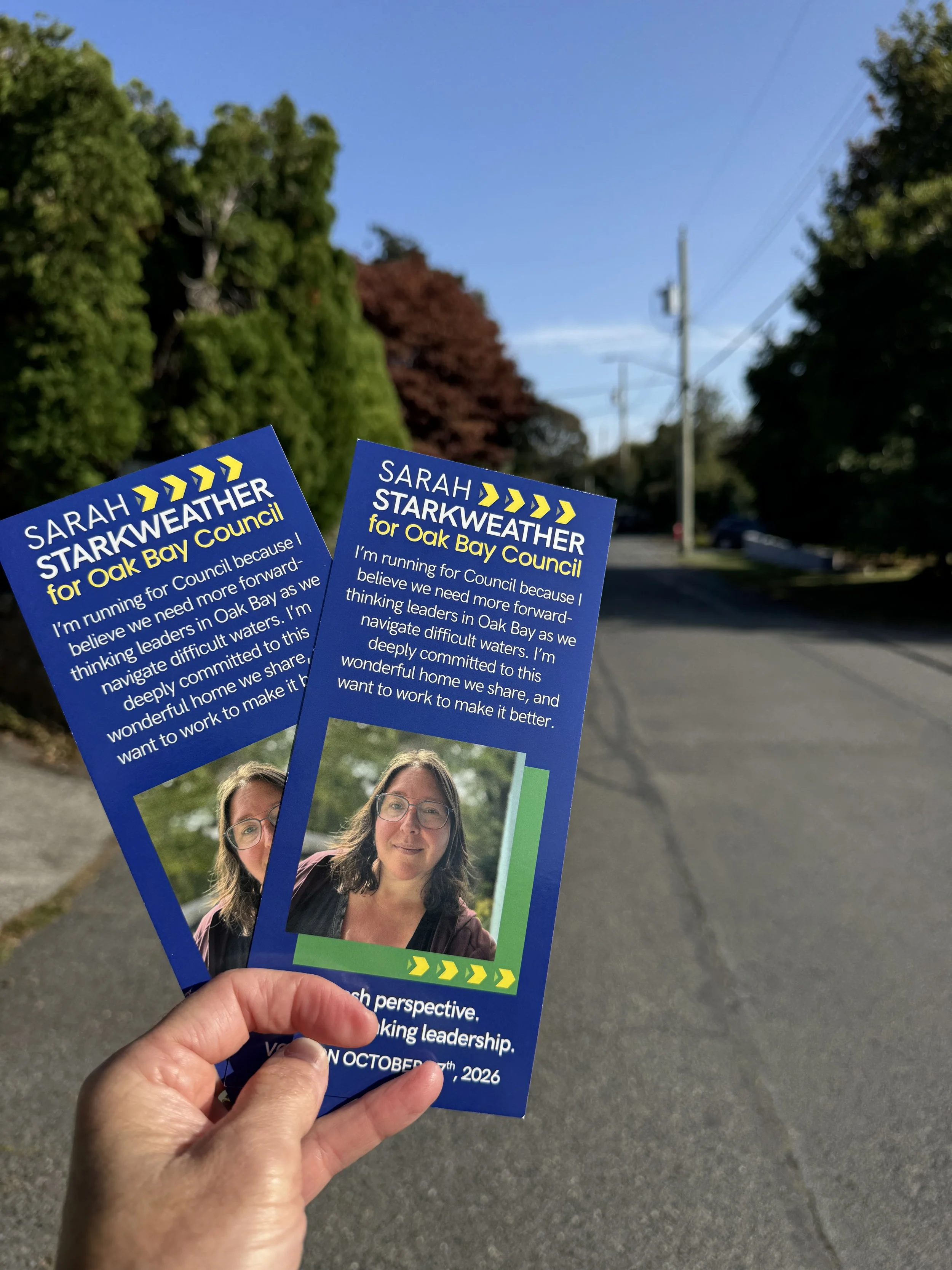 A person holding two campaign flyers for Sarah Starkweather for Oak Bay Council, with a background of a street lined with trees and utility poles on a clear day.