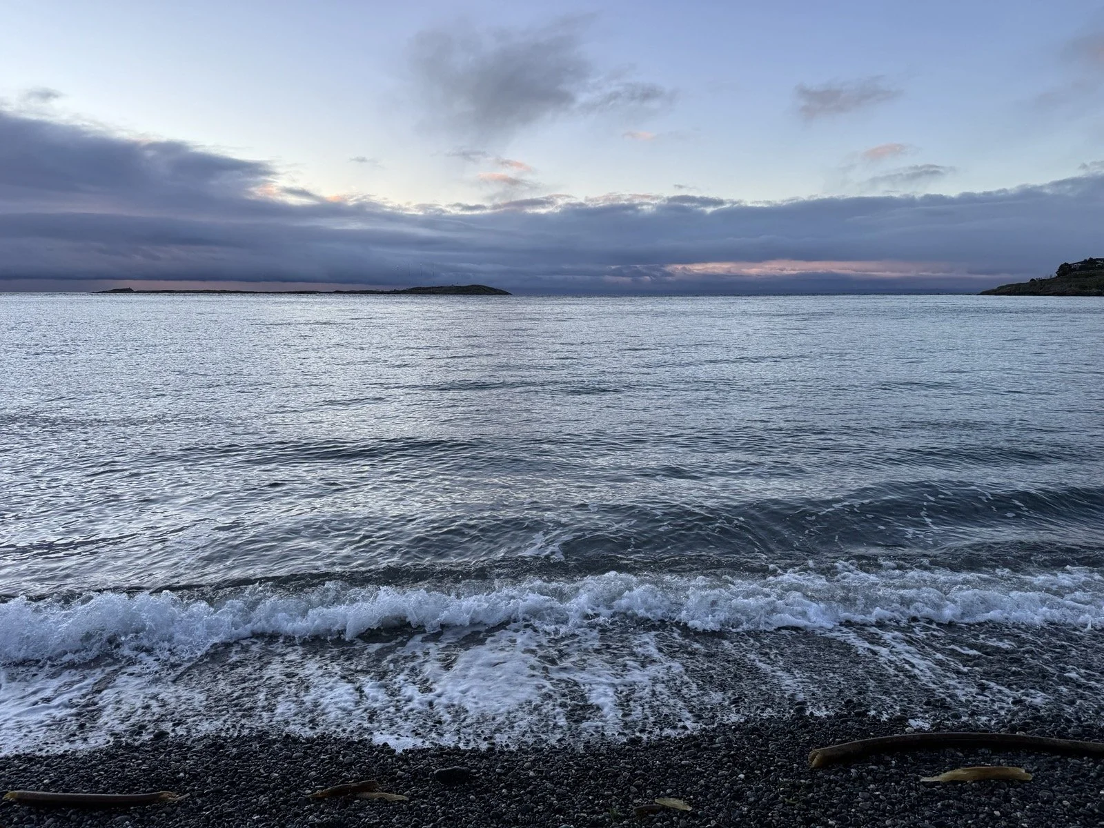 A calm ocean view with a cloudy sky, distant land mass, and gentle waves lapping a pebbled shore.