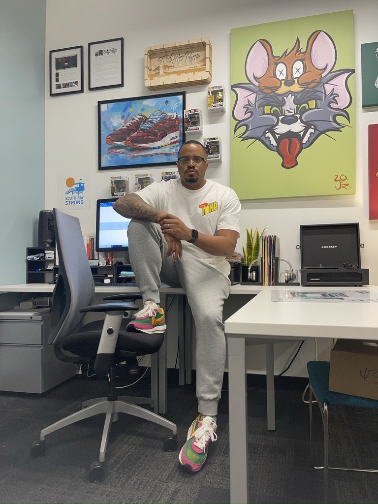 A man sitting on a black office chair in an office space, wearing a white T-shirt, grey sweatpants, and colorful Nike sneakers. The wall behind him features various framed artwork, including a large colorful cartoon cat with a mouse on its head, and a painting of sneakers.