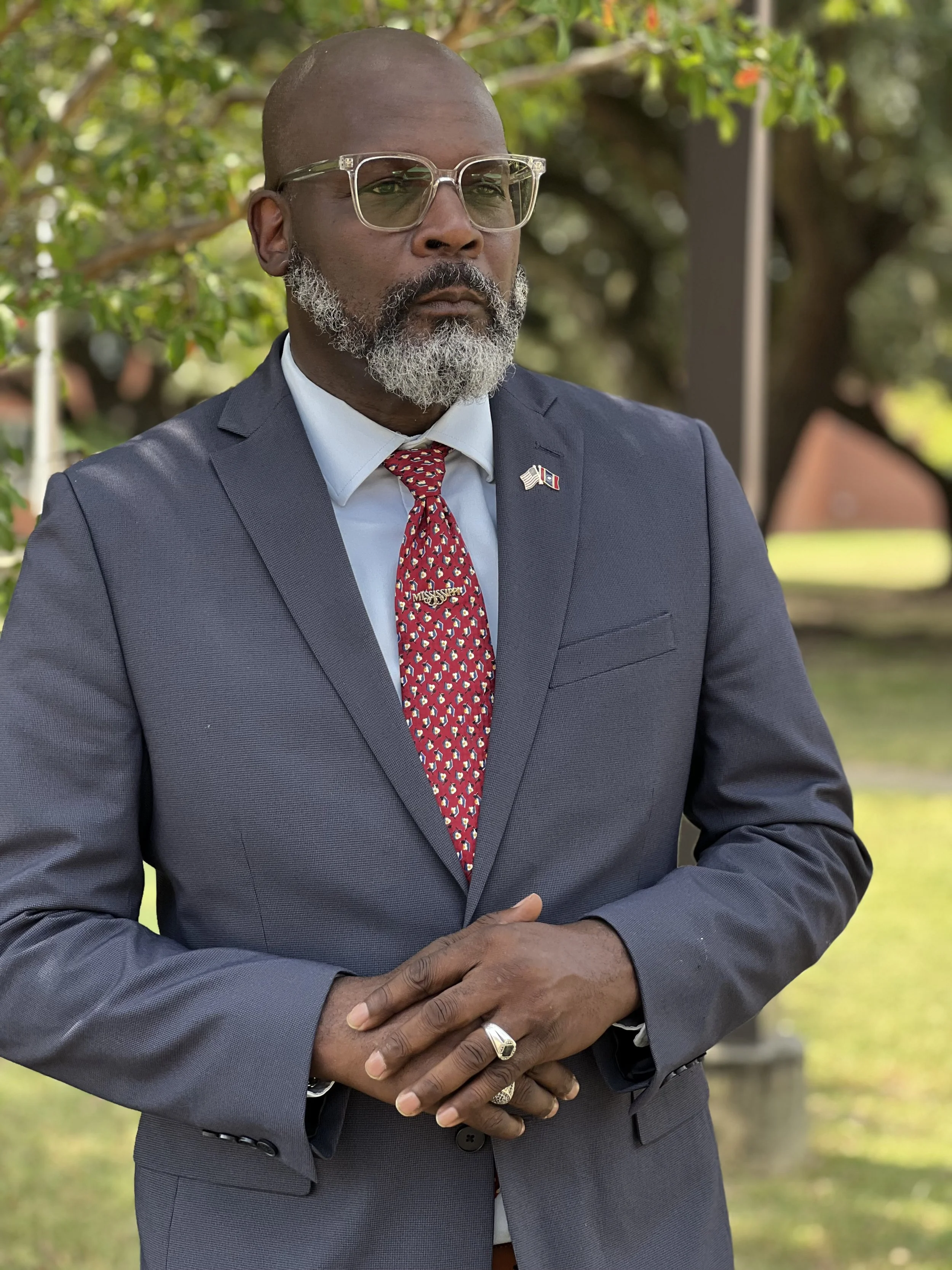A man in a gray suit with a red patterned tie and American flag lapel pin, wearing glasses and a ring, standing outdoors with trees and blurred background.