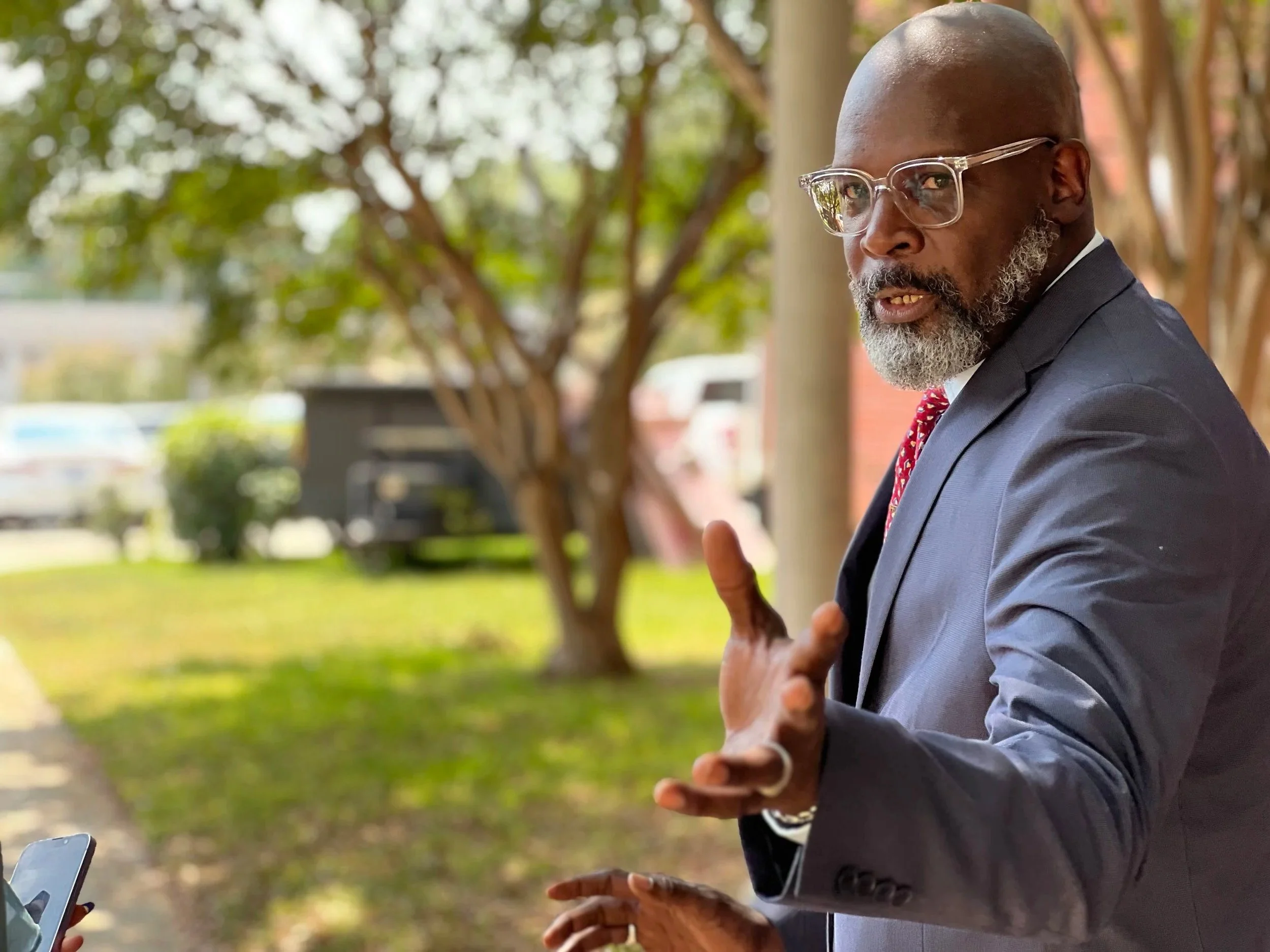 A man with a bald head and gray beard, wearing glasses and a dark suit with a red patterned tie, is outdoors, seemingly talking or explaining something, with trees and a building in the background.
