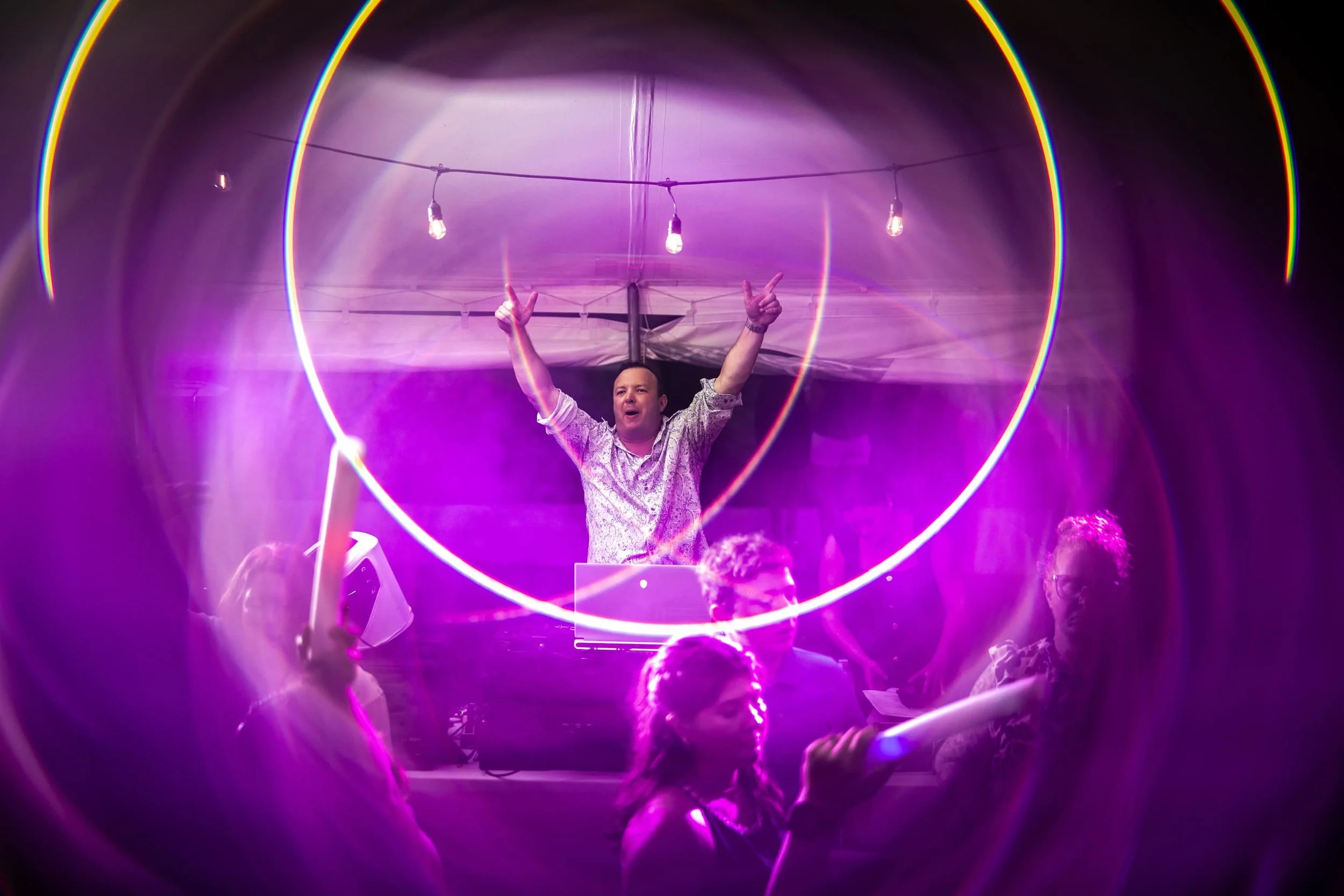 DJ raising both arms above the booth as wedding guests dance under purple lighting and string lights.