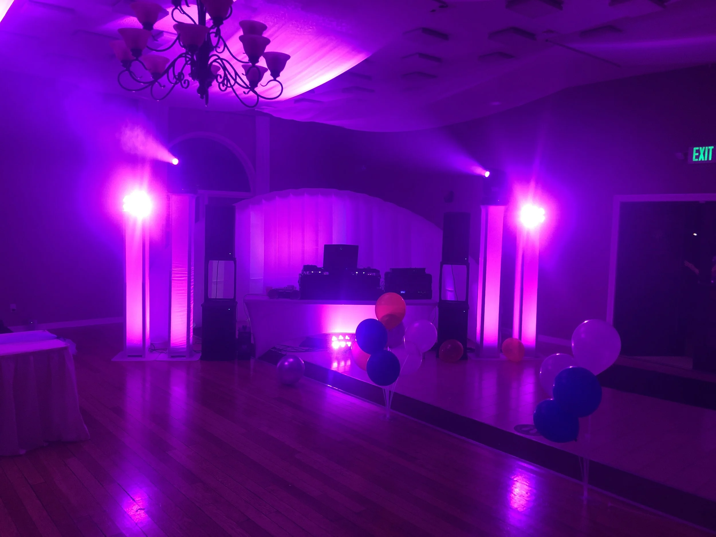 Purple light beams fill a balloon-decorated dance floor in front of a DJ booth.
