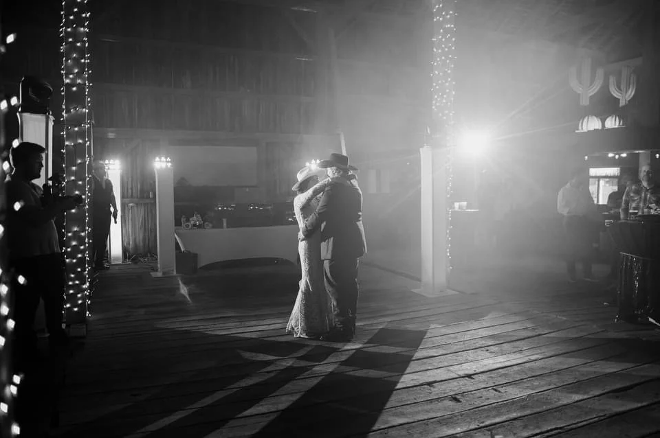 Black and white photo of a couple slow dancing in a barn reception space.
