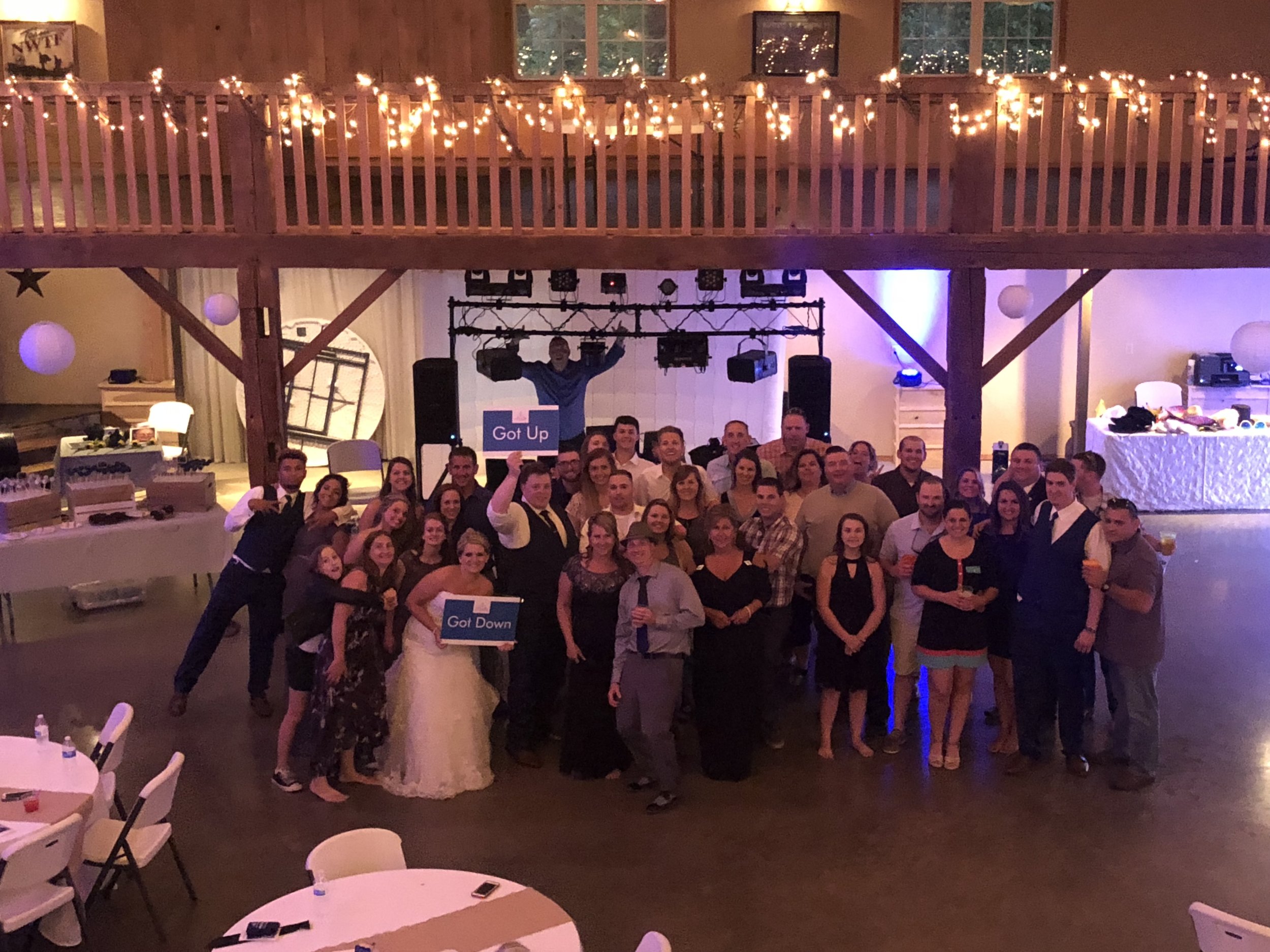 Wedding guests gathered in a barn reception hall with string lights and a DJ setup behind them.