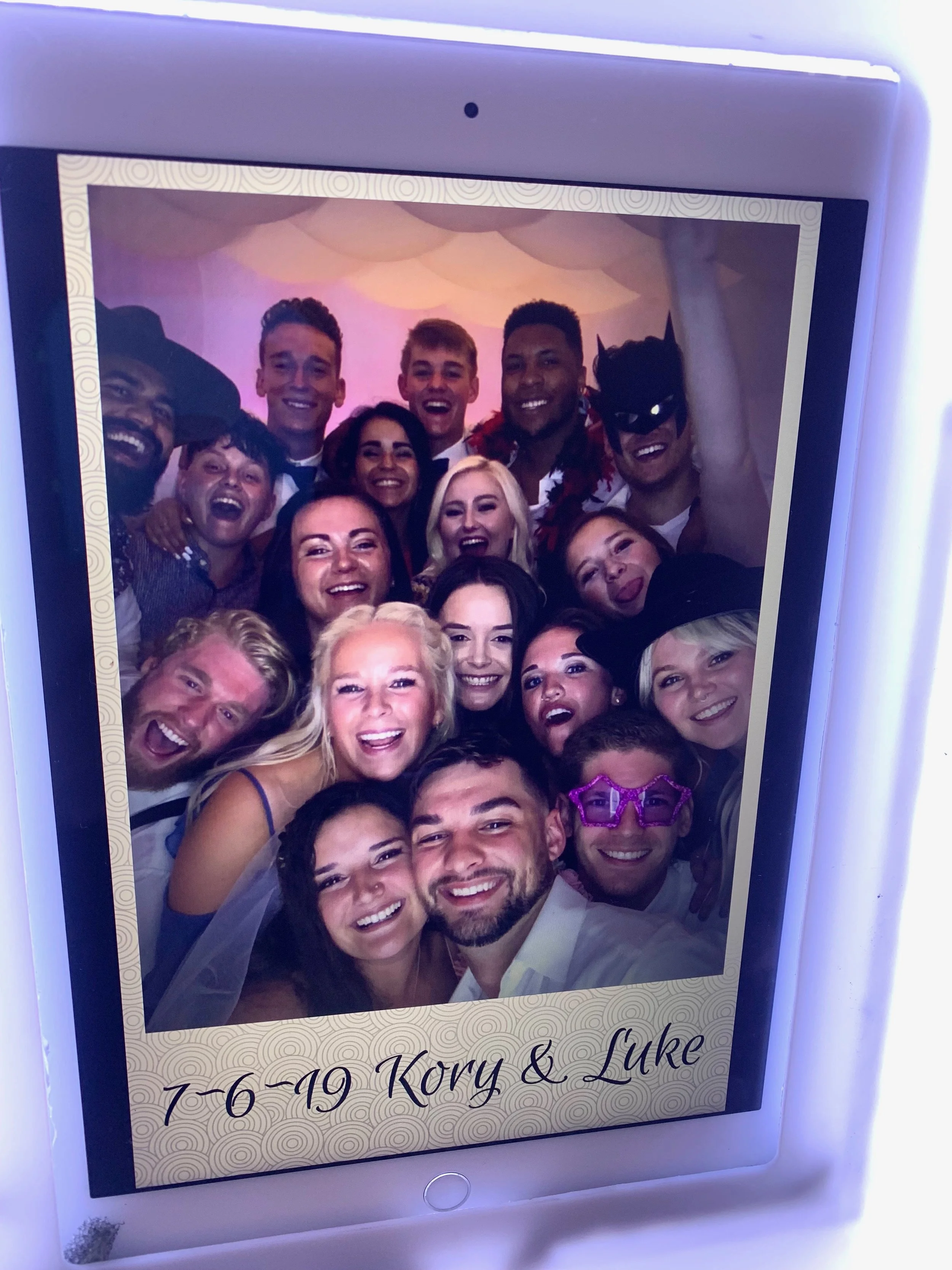 Photo booth tablet displaying a smiling group wearing playful hats, masks, and glasses.