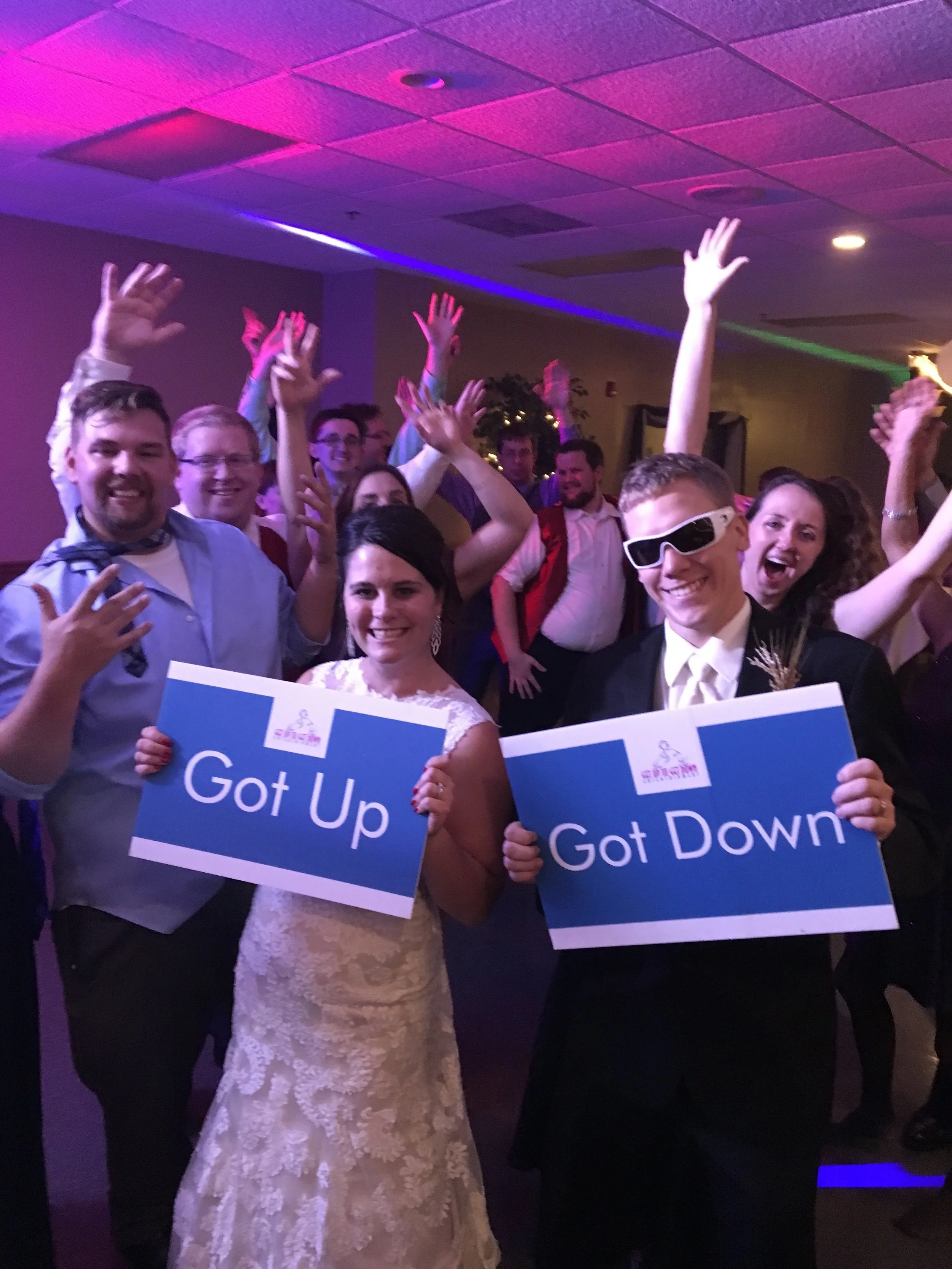 Bride, groom, and guests holding Got Up and Got Down signs under pink reception lighting.