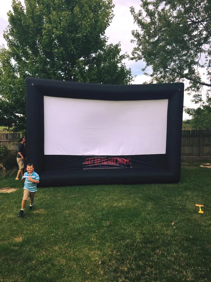Large inflatable movie screen set up on a lawn beside trees.