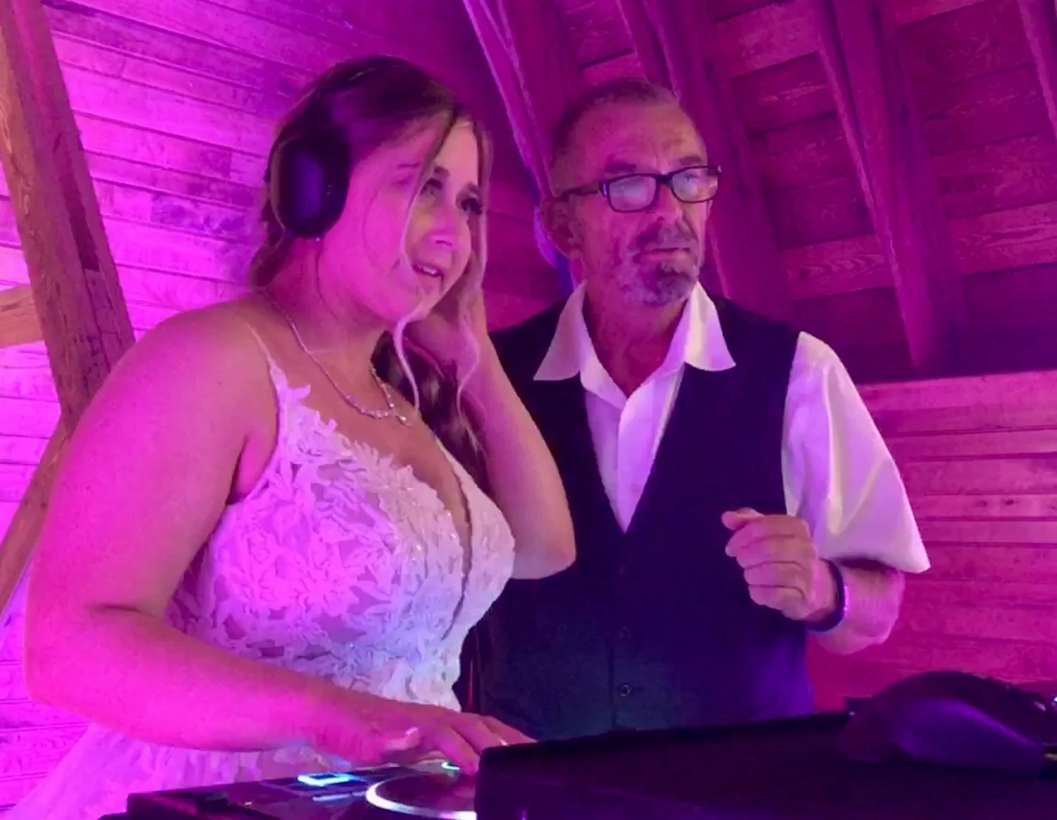 Bride wearing headphones at a DJ controller beside a man in a vest under pink lighting.