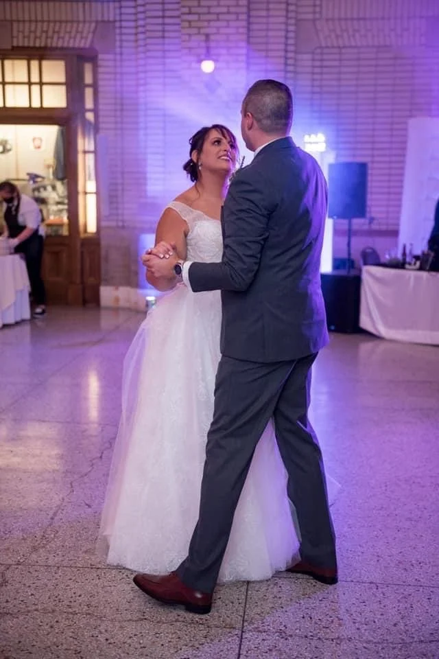 Bride dancing with a man in a dark suit beside purple reception lighting.